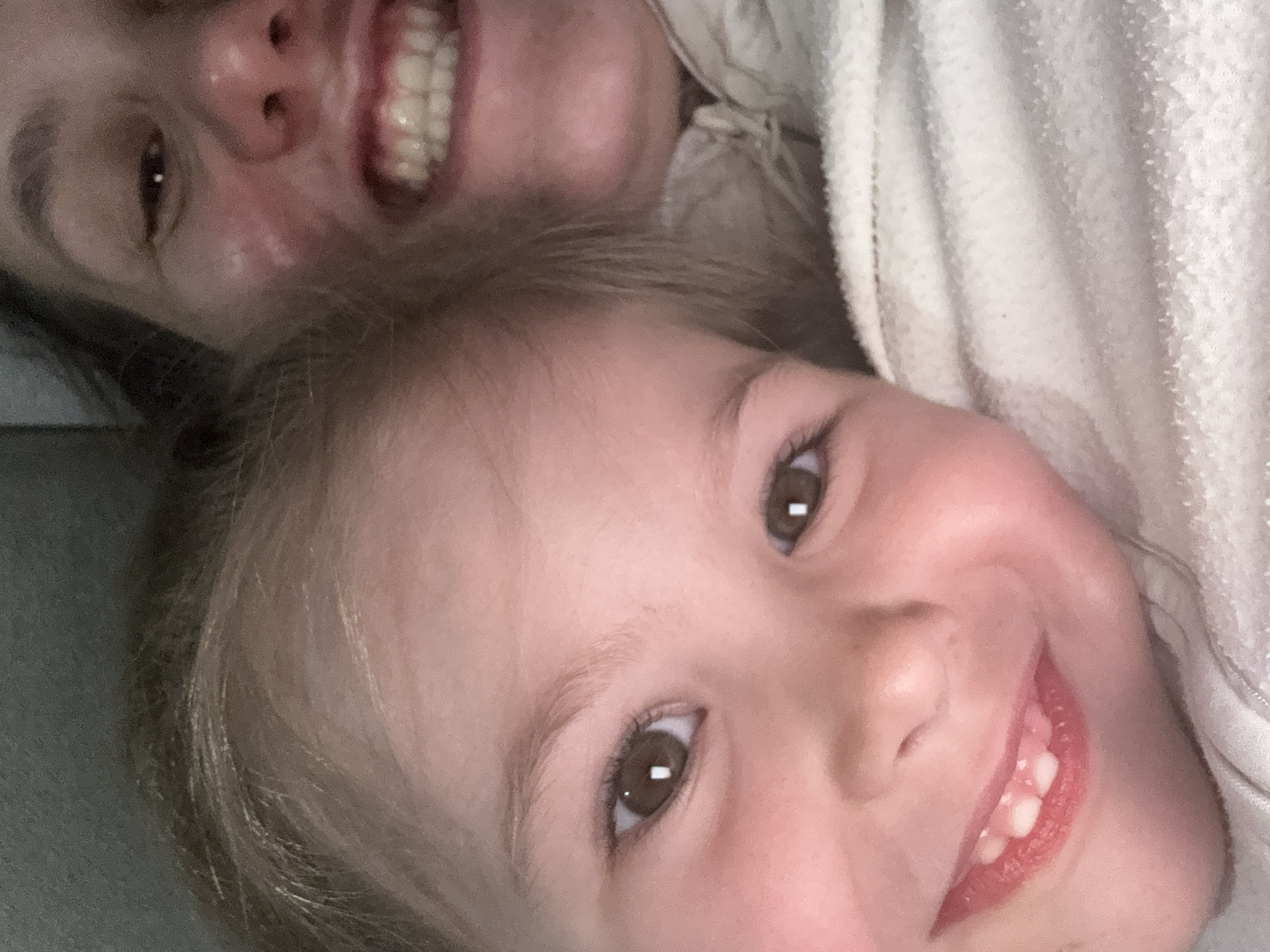 The image depicts two individuals, presumably a mother and daughter, closely positioned together. The child is in the foreground, with a wide smile revealing teeth. The adult is behind the child and also smiling. The background consists of a soft, light-colored blanket or fabric. The image is slightly rotated. There is no evidence to confirm or suggest that the picture was taken in the city of Zelenogradsk, Russia.