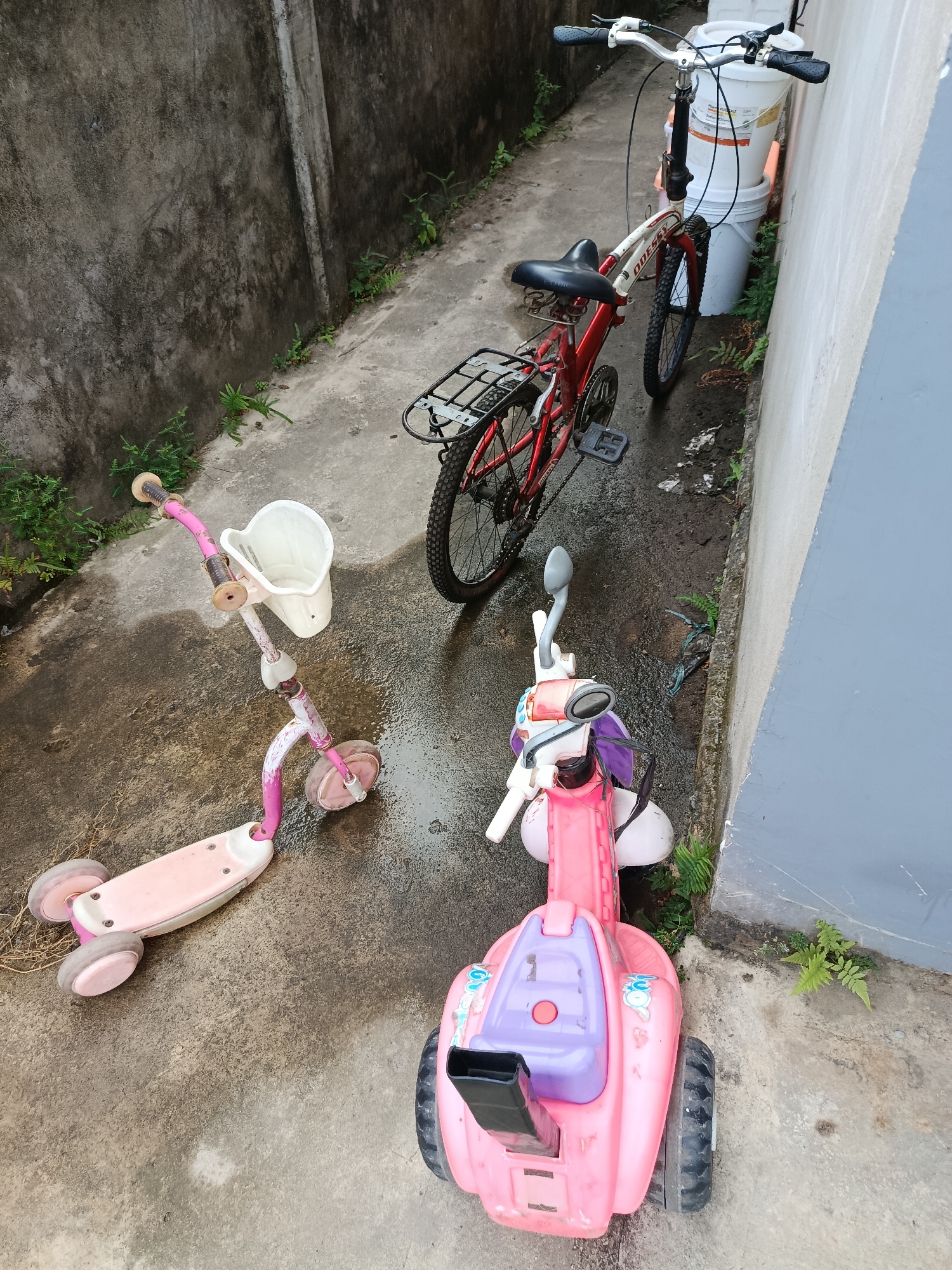 This image depicts a narrow outdoor alleyway with a concrete floor and two walls. One wall is rough and grey, while the other is smooth and light grey. The alley is wet, suggesting recent rain. Several children's toys are present: a red and white bicycle, a pink scooter with a white basket, and a pink ride-on toy. The bicycle is positioned in the middle of the alley. The scooter is to the left of the bicycle, and the pink ride-on toy is to the right. There are no people visible in the image. The scene appears to be a residential area in Palembang, Indonesia, likely a backyard or side yard.