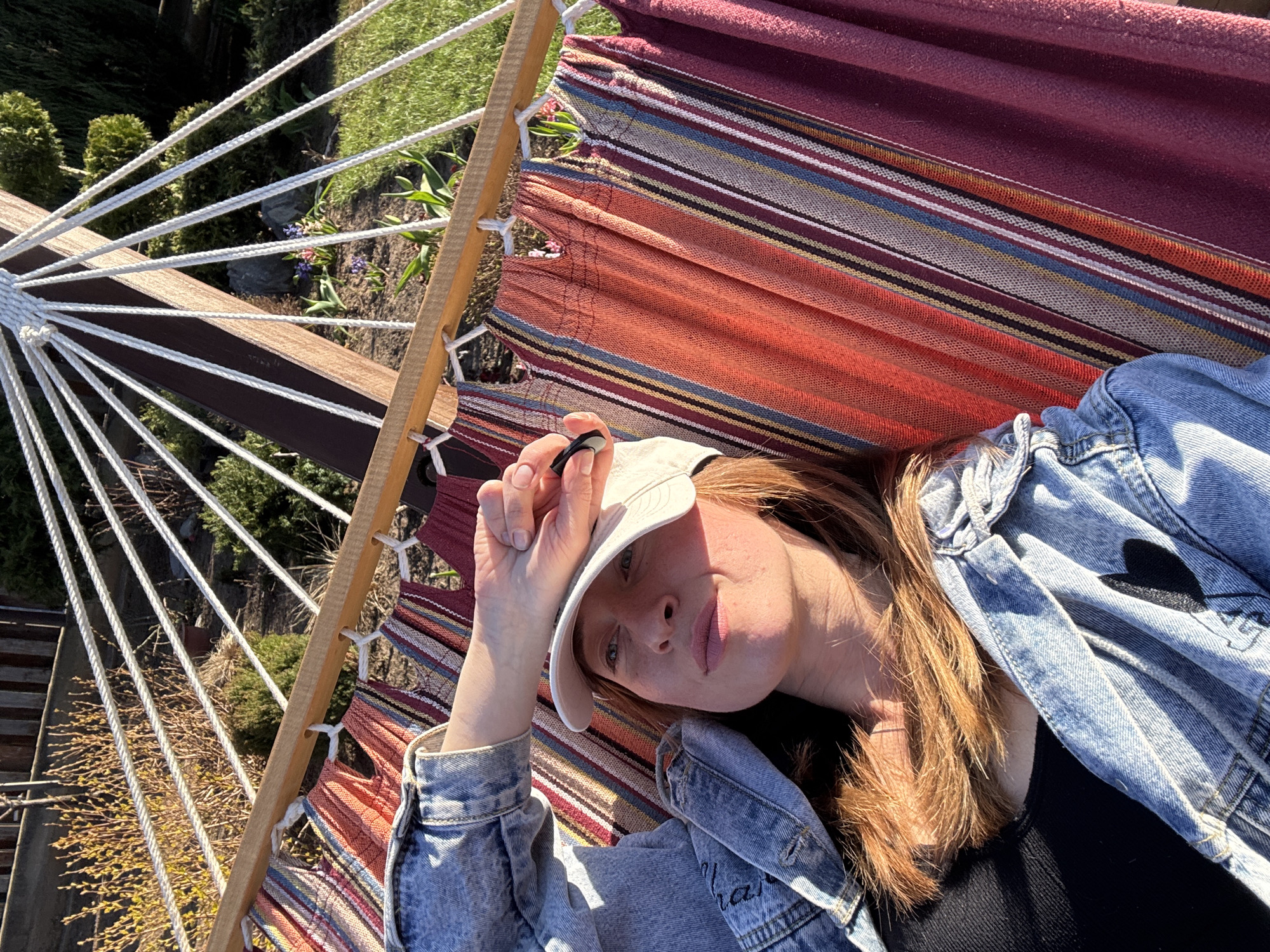 A person lies in a hammock, wearing a denim jacket and a baseball cap. The hammock is striped with shades of red, orange, and blue. The background shows a garden with green bushes and a fence. The location is unknown and cannot be determined from the image.