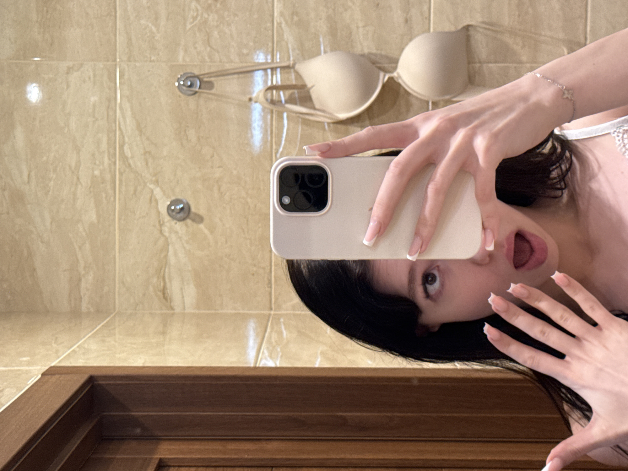 A young woman with dark hair takes a selfie in what appears to be a bathroom. She is holding a smartphone in her right hand, and her left hand is held up with fingers spread. Her mouth is open in a surprised expression, and her eyes are wide. Her nails are long and painted with a French manicure. She is wearing a white lace top. In the background, a beige bra is hanging on a hook against a tiled wall. To the left, a wooden doorframe is visible. The overall setting suggests a private residential space. The image has been identified as being taken in Khimki, Russia.