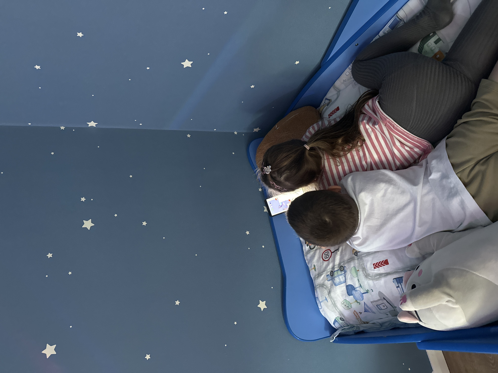 Two children are lying in a blue bed, looking at a mobile device. One child wears grey leggings and a red and white striped shirt, and the other wears a white shirt and khaki pants. A large white plush animal is also in the bed. The room has dark blue walls with white star decorations. The location is the city of Chaikovsky, Russia.