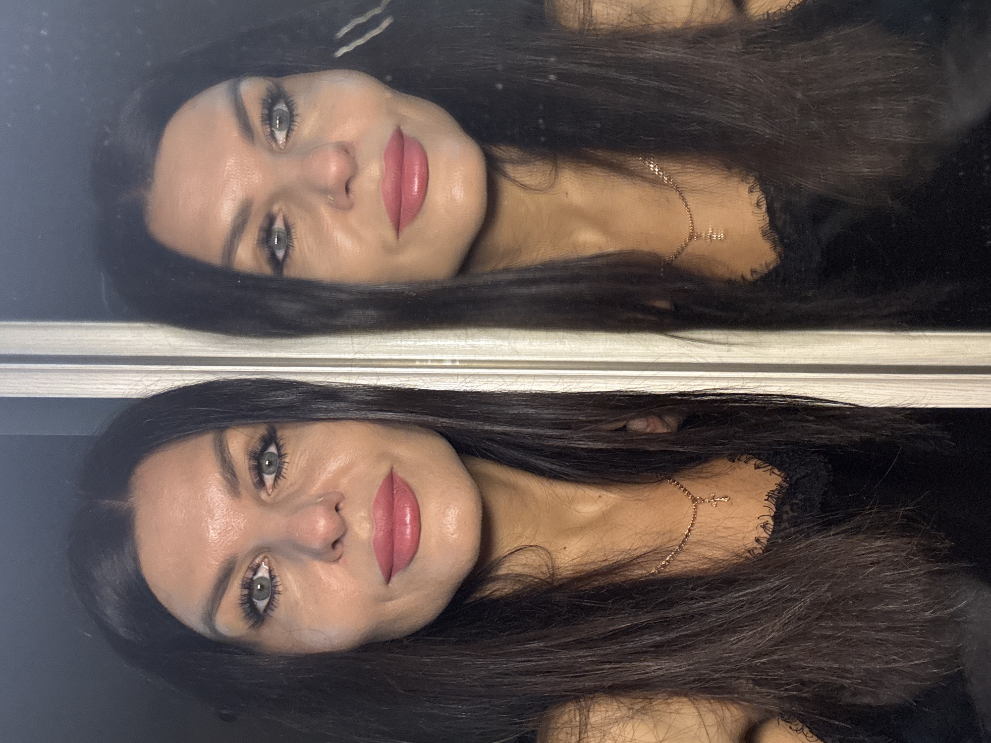 A woman is reflected in a mirror. She has dark hair, light eyes, and pink lipstick. She is wearing a gold necklace with a cross pendant. The location is not discernible from the image.