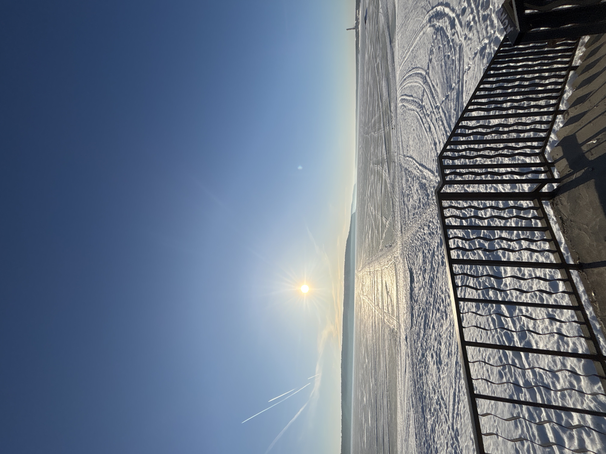 This photo, taken in Chaikovsky, Russia, shows a winter landscape with clear blue skies and a bright sun. The foreground features a metal staircase with its shadow cast on the snow-covered ground. The ground is covered in snow, with various tracks suggesting prior activity. Jet contrails are visible in the sky. The horizon line appears to delineate a snow-covered landscape or frozen body of water stretching towards the distant shore. The photograph provides a wide perspective, showcasing the natural environment and elements of the city in a wintry setting.