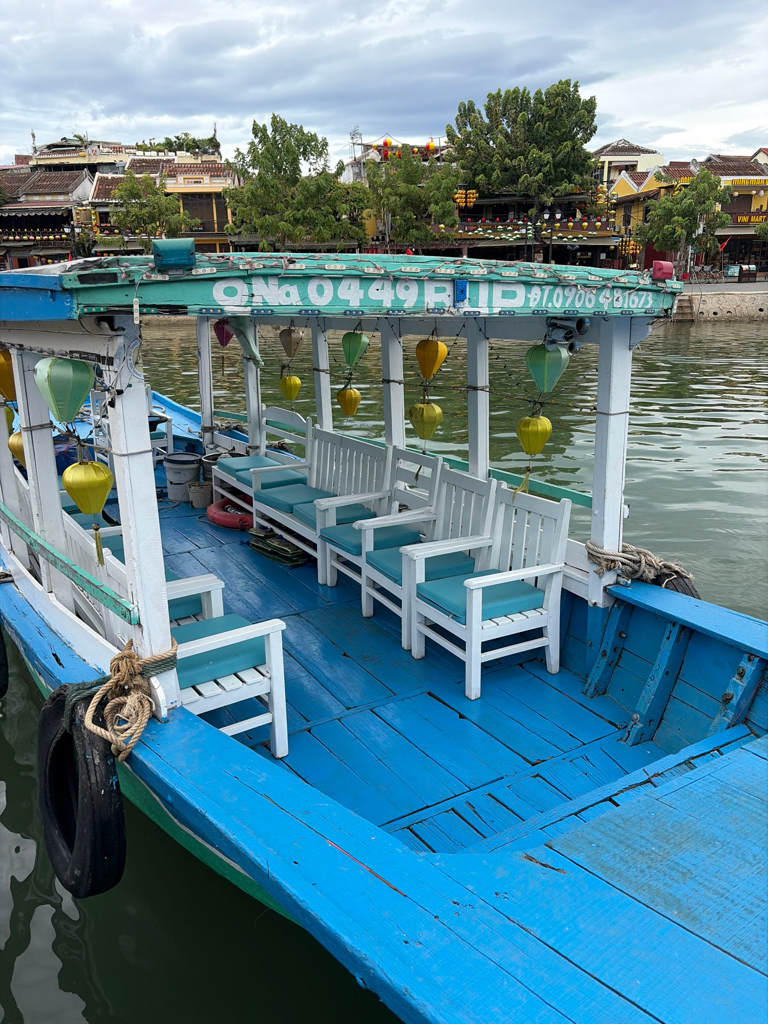 The image depicts a blue wooden boat docked on a calm body of water, likely a river or canal in Hoi An, Vietnam, given the architectural style of the buildings visible in the background. The boat is equipped with several white wooden chairs with teal cushions, arranged in rows. Decorative lanterns, primarily yellow and green, hang from the boat's canopy, which is painted teal and white and displays signage with the text "ONG 0449.RIP Dt.0906 481673". A black tire is secured to the side of the boat with rope, acting as a fender. The water is calm with ripples, reflecting the overcast sky. The background features traditional Hoi An buildings with tiled roofs and vibrant yellow and orange facades, interspersed with lush green trees and foliage. The scene is devoid of people, suggesting the boat is either awaiting passengers or has just completed a tour.