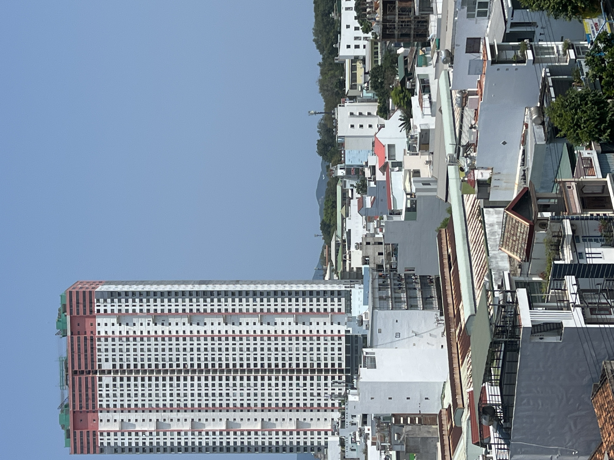 The image depicts an urban scene in Nha Trang, Vietnam. Dominating the left side of the frame is a tall, unfinished high-rise building, characterized by its repetitive window patterns, pink/red horizontal stripes, and exposed concrete structure. The surrounding urban landscape includes densely packed buildings, primarily low-rise and medium-rise, interspersed with greenery. Building exteriors appear primarily white. The scene is set under a clear, light-blue sky.