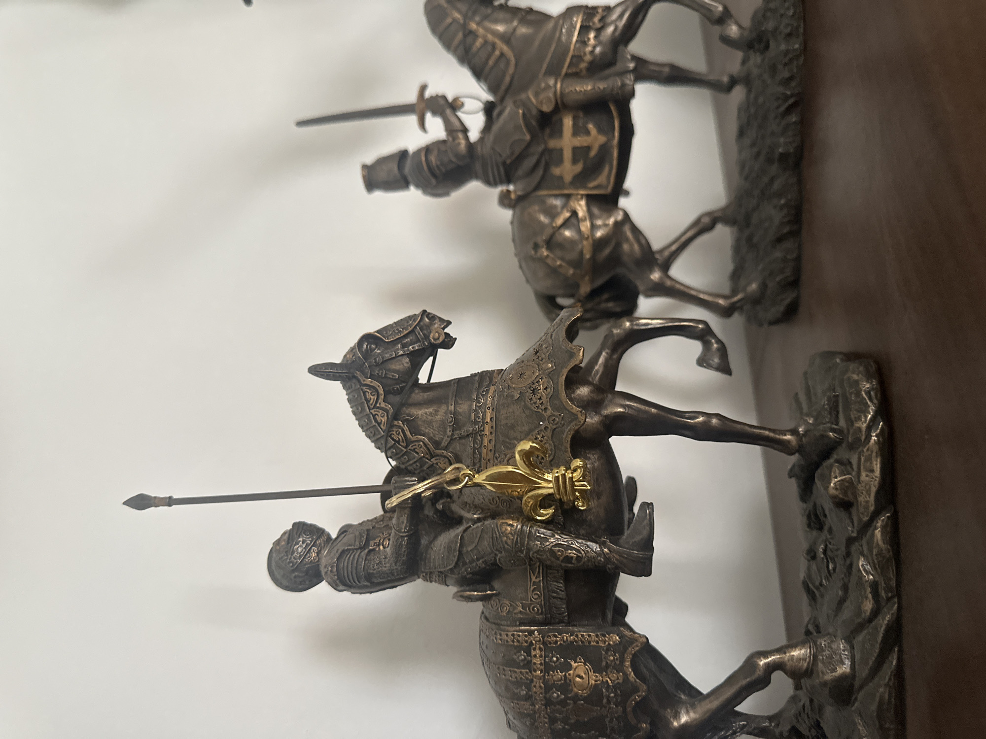 The image depicts two bronze statues of medieval knights on horseback. The knights are armored and carry weapons. The bottom statue has a golden fleur-de-lis attached. The statues appear to be indoors against a white wall and a dark brown surface. There is no information to indicate that the image was taken in Sofia, Bulgaria.