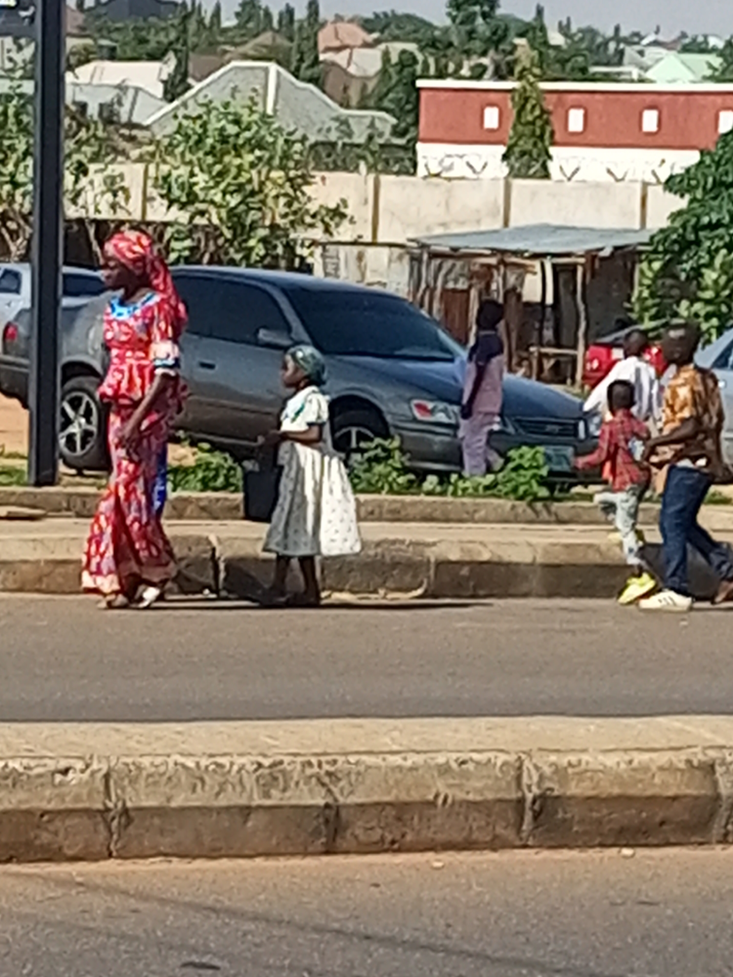 In Tumfure, Nigeria, a group of people are walking along a street. In the foreground, a woman in a red and white patterned dress and headscarf walks from left to right. Beside her, a young girl in a white dress walks in the same direction. Behind them, a dark gray sedan is parked on the side of the road. Further back, other individuals can be seen near a building with a red and white facade. The environment appears to be a dry, urban setting with trees and buildings visible in the background. The road has a concrete curb separating it from the sidewalk area where the pedestrians are walking.