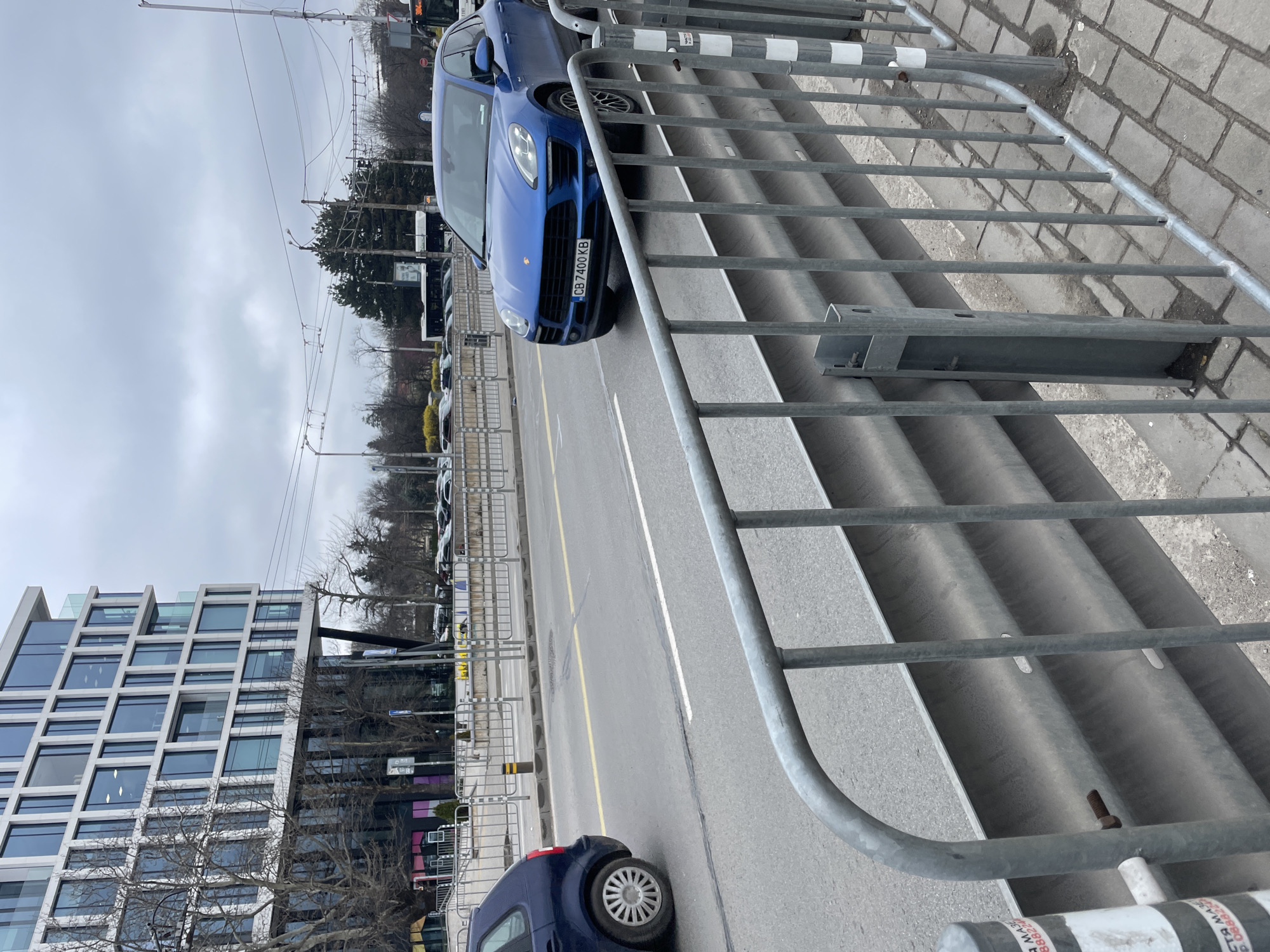 The image captures an urban scene in Sofia, Bulgaria, featuring a street with cars and a modern building. A blue Porsche Cayenne is partially visible in the foreground, its license plate reading "CB 7400 KB". Behind it, several other vehicles are parked or moving along the street. A contemporary building with a grid-like facade stands on the left side of the frame. A metal barrier partially obscures the view, with its bars running diagonally across the scene. Overhead power lines and poles are visible against a cloudy sky. The road has a yellow centerline, and a white line designates the lane's edge.