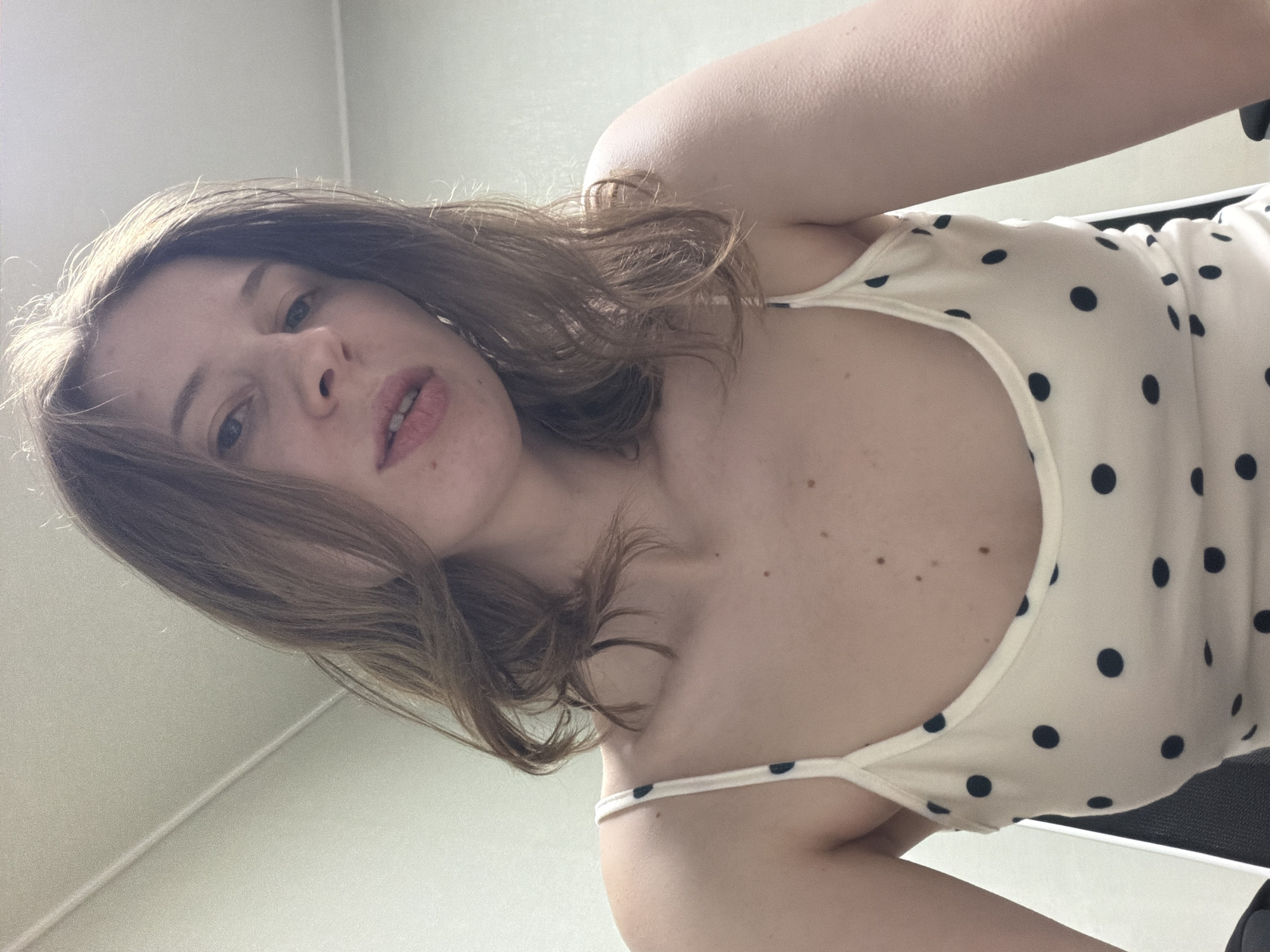 A young woman with shoulder-length brown hair is shown from the chest up. She is wearing a white tank top with navy blue polka dots. The background appears to be an indoor setting with a light-colored wall and a ceiling fixture. The lighting is soft and diffused. There is no indication of the city of Dongducheon-si, South Korea, in the image.