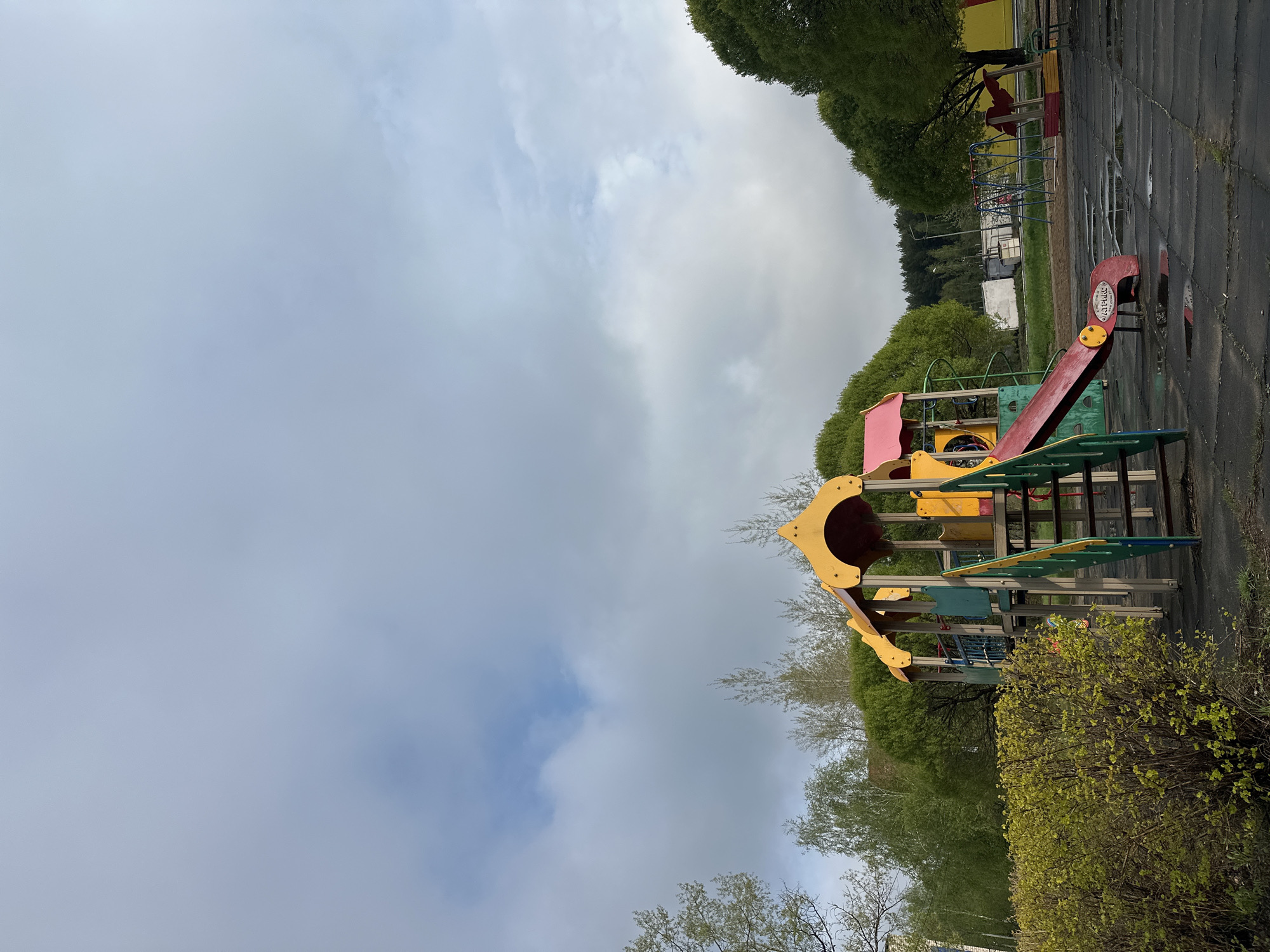 The image shows a playground in the city of Chaikovsky, Russia. The playground features colorful play structures including slides, climbing frames, and other equipment. The surrounding environment includes trees, bushes, and some buildings in the background. The sky is overcast.