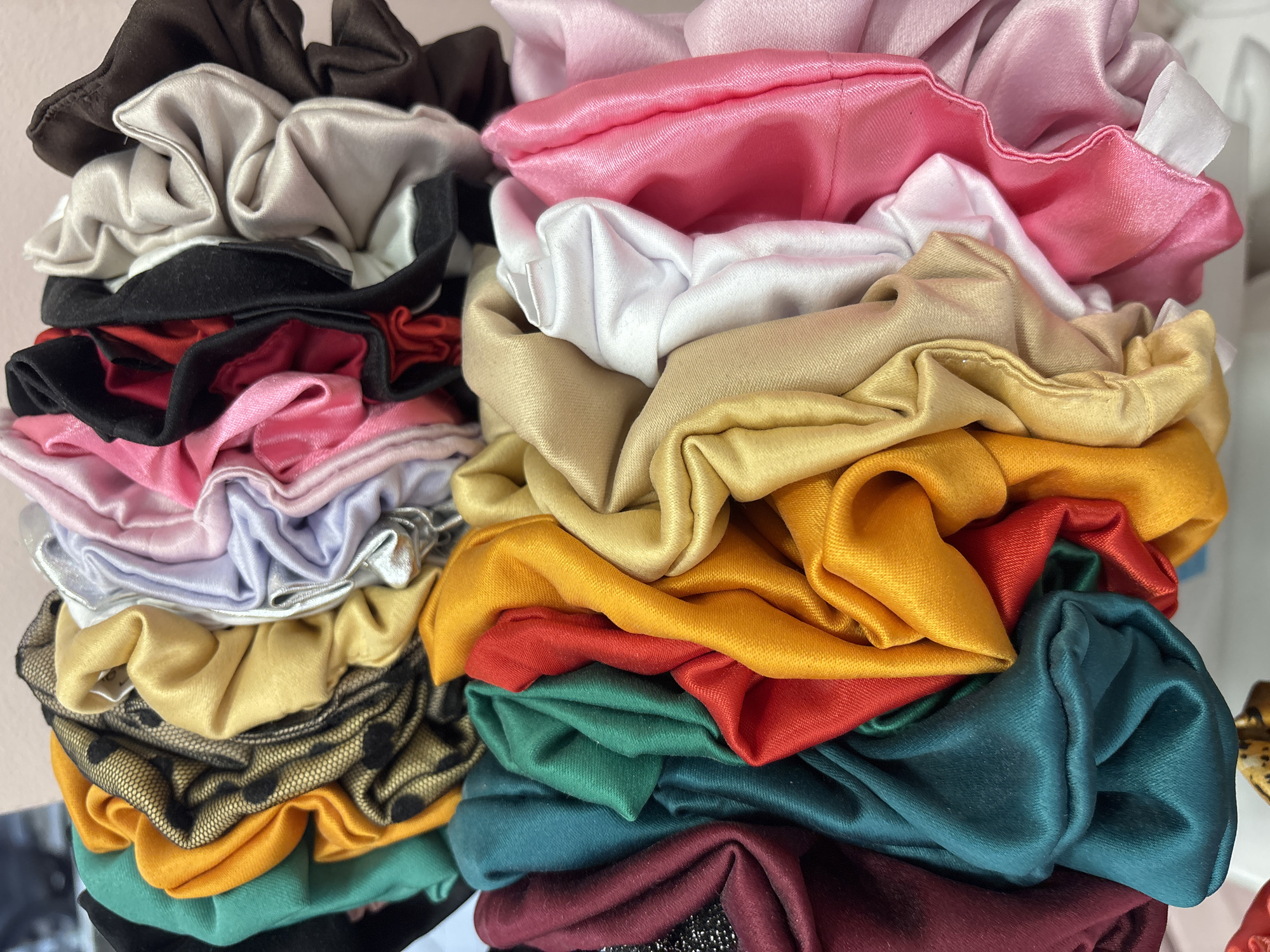 The image is a close-up, eye-level shot of a large, vibrant collection of scrunchies stacked in two main piles. The scrunchies exhibit a wide array of colors and textures, with many appearing to be made of satin or silk-like fabric due to their lustrous sheen.
The left stack features scrunchies in colors such as dark brown, silver-grey, black, bright red, various shades of pink, light purple, light blue, and a golden-yellow. Notably, a black mesh scrunchie with black polka dots is visible in this stack, layered over other fabrics.
The right stack presents a more gradual color progression, starting with soft pinks, white, and pale gold at the top. This transitions into richer tones of yellow, orange, red, emerald green, deep teal, and burgundy towards the bottom.
The scrunchies are arranged in a dense, slightly haphazard manner, highlighting their ruffled and gathered forms. The lighting is soft and even, emphasizing the fabric's folds and reflective qualities. No specific activity or event is taking place; the scrunchies are simply displayed. There are no people, discernible location cues related to Sofia, Bulgaria, or visible text within the frame.