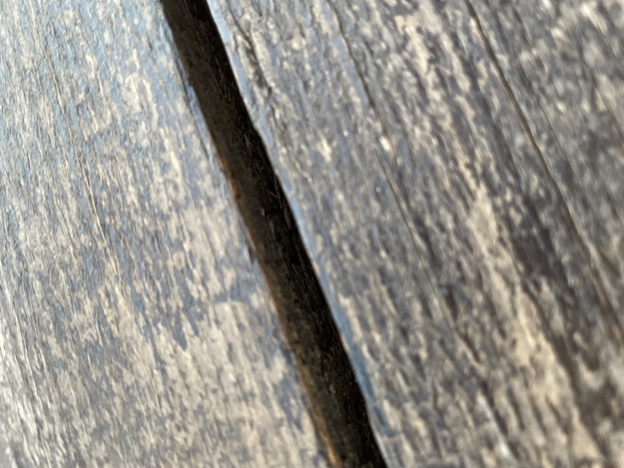 The image is a close-up, abstract view of weathered wooden planks. The planks are dark gray and have a rough, textured surface with visible grain patterns and imperfections. There is a prominent dark, deep crack or seam running diagonally across the center of the frame, separating two planks. The lighting suggests it is daytime, with highlights and shadows emphasizing the texture and contours of the wood. The overall impression is one of age and natural wear. There is no discernible text, people, or specific scene depicted, making the subject purely textural and focused on the material.