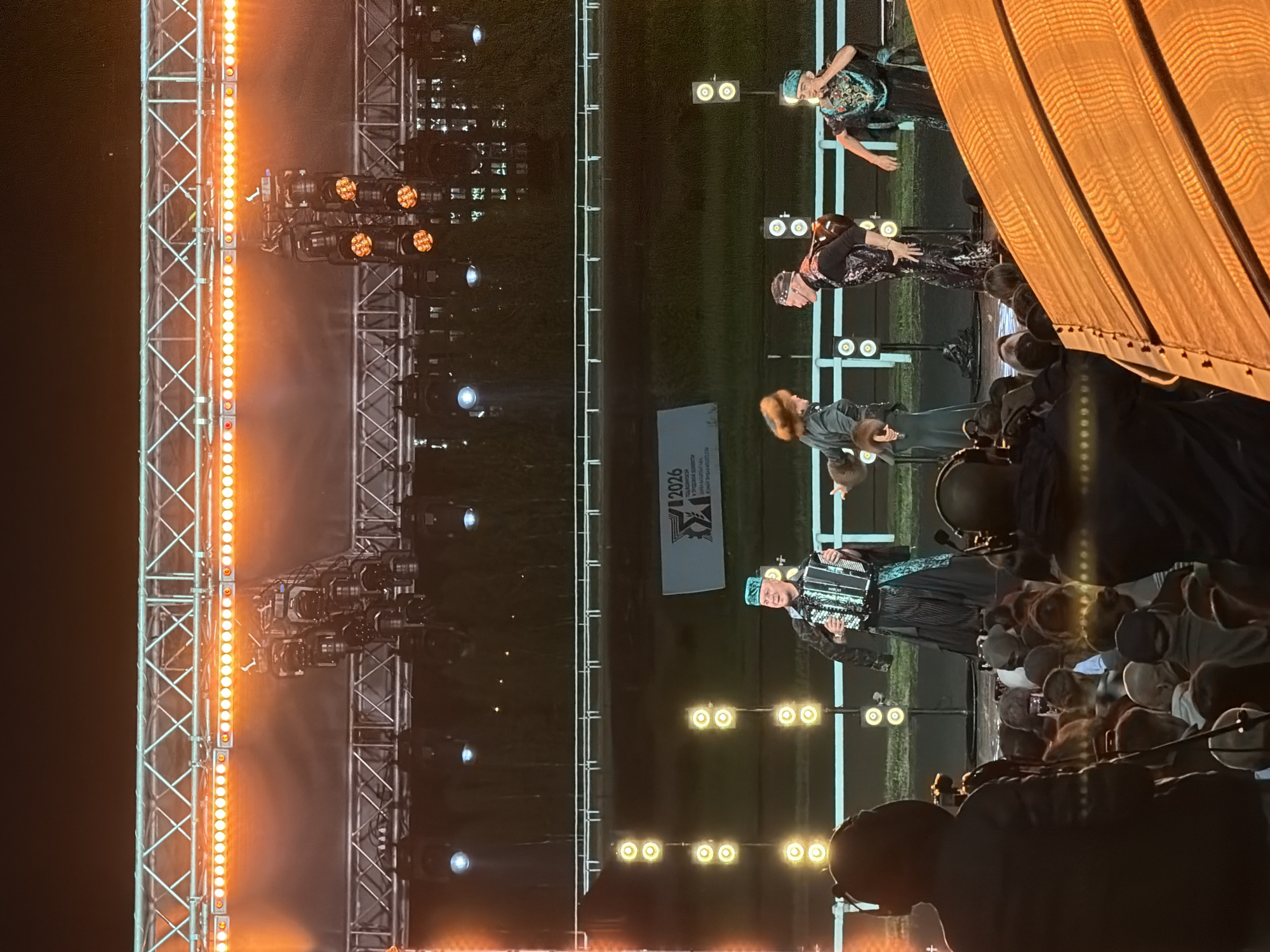 An outdoor nighttime performance is underway. The stage is lit with bright orange lights, and a large metal truss system supports various lighting fixtures. A banner with "2026" and an emblem is visible behind the performers. In the foreground, a crowd of people watches the show. On stage, several performers are dressed in traditional or elaborate costumes. One performer on the left is playing an accordion, wearing a traditional hat and a black coat with ornate trim over a striped shirt. To the right of the accordion player, a female performer with bright red hair wears a dark coat and fur accessories. Another male performer, in the center, wears a black vest with a patterned design over a black shirt and headband. To the far right, a performer in a vibrant, patterned costume and a turquoise hat sings into a microphone. The background consists of a dark field and a white railing.