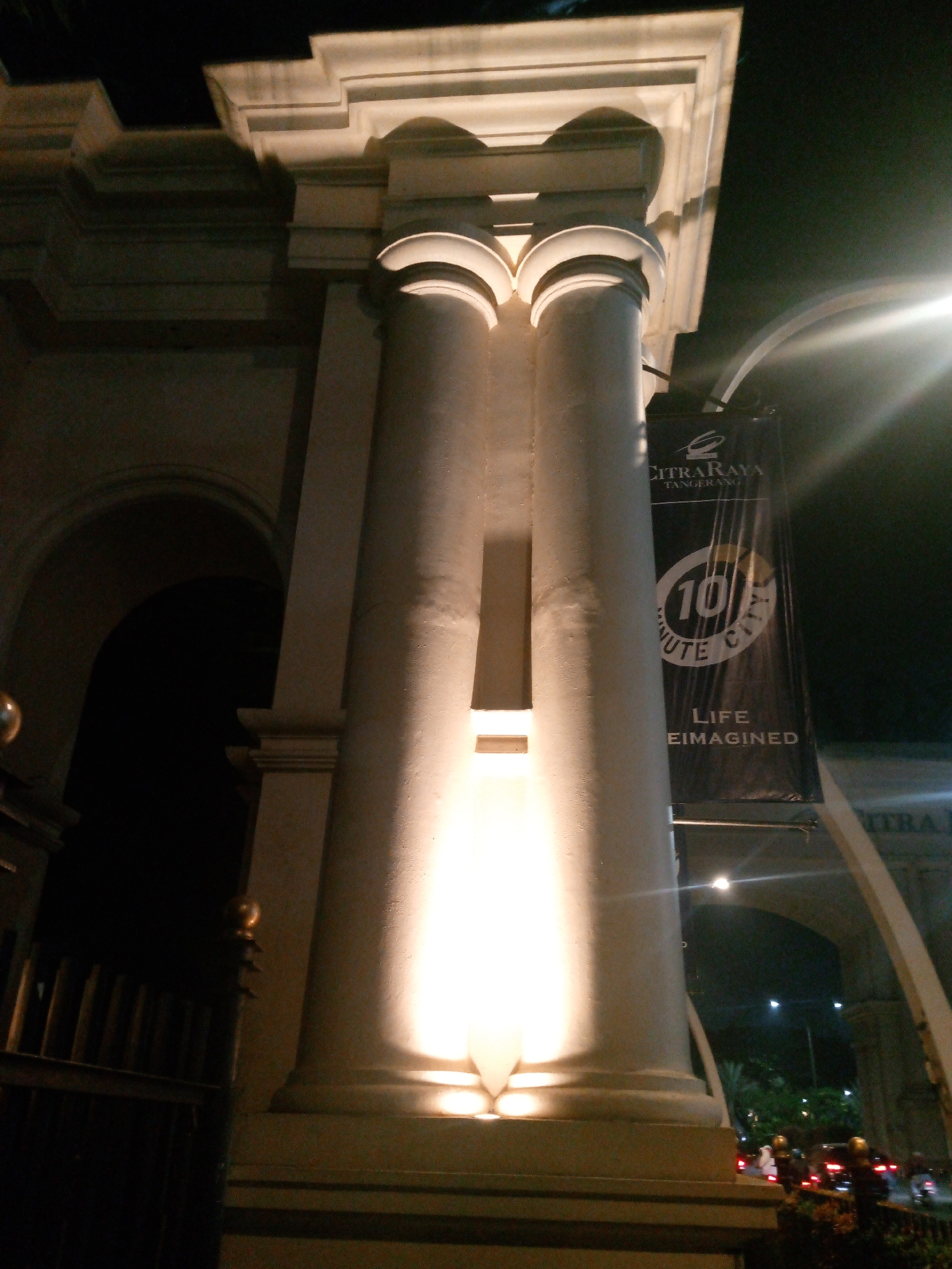 An eye-level, wide shot captures a dimly lit, nighttime view of an ornate architectural structure with two large, fluted columns illuminated by spotlights. The columns are capped by a decorative entablature. A dark banner hangs between the columns, displaying text that reads "CITRARAYA TANGERANG" and a circular logo with "10 MINUTE CITY" and "LIFE REIMAGINED." The background is dark, with glimpses of an archway, streetlights, and vehicular traffic. The overall scene suggests an entrance or gateway to a developed area, possibly a commercial or residential complex. The image is taken at night, with artificial lighting highlighting the architectural features and the banner. The location is specified as Bitung Jaya, Indonesia.