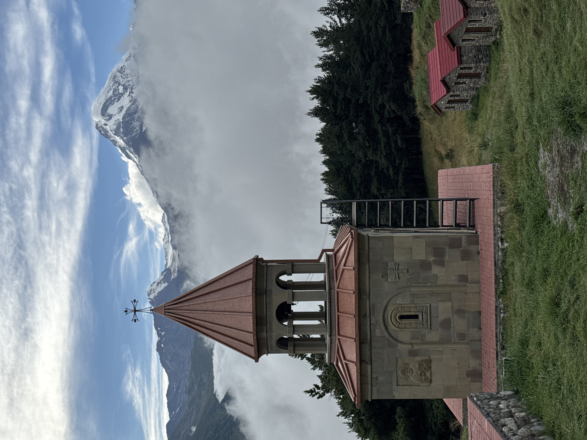 A stone church with a red copper roof stands on a grassy hill. The church has a bell tower with arched openings and a cross on top. To the right of the church, there are three small stone houses with red roofs. In the background, a majestic snow-capped mountain rises above a sea of clouds. The sky is blue with scattered white clouds. The image appears to be taken in Stepantsminda, Georgia, a town known for its stunning mountain scenery.