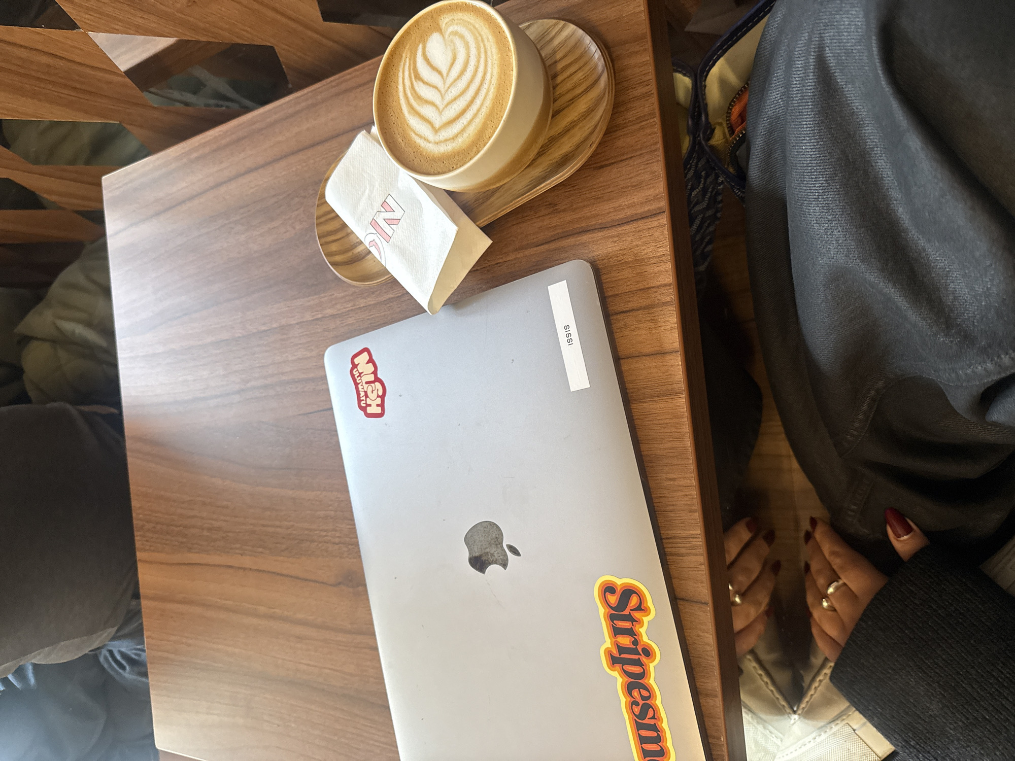 A latte with latte art sits on a small wooden table alongside a laptop. The laptop has two stickers. One sticker says "Stripesman" in an orange and yellow font, the other sticker is partially obscured, but contains the word "Kush". A napkin sits next to the coffee. The table is wood, and the person is wearing dark clothing and has rings on their fingers and red nail polish. The setting appears to be a cafe. It is impossible to determine the specific location within Paris from the image alone.