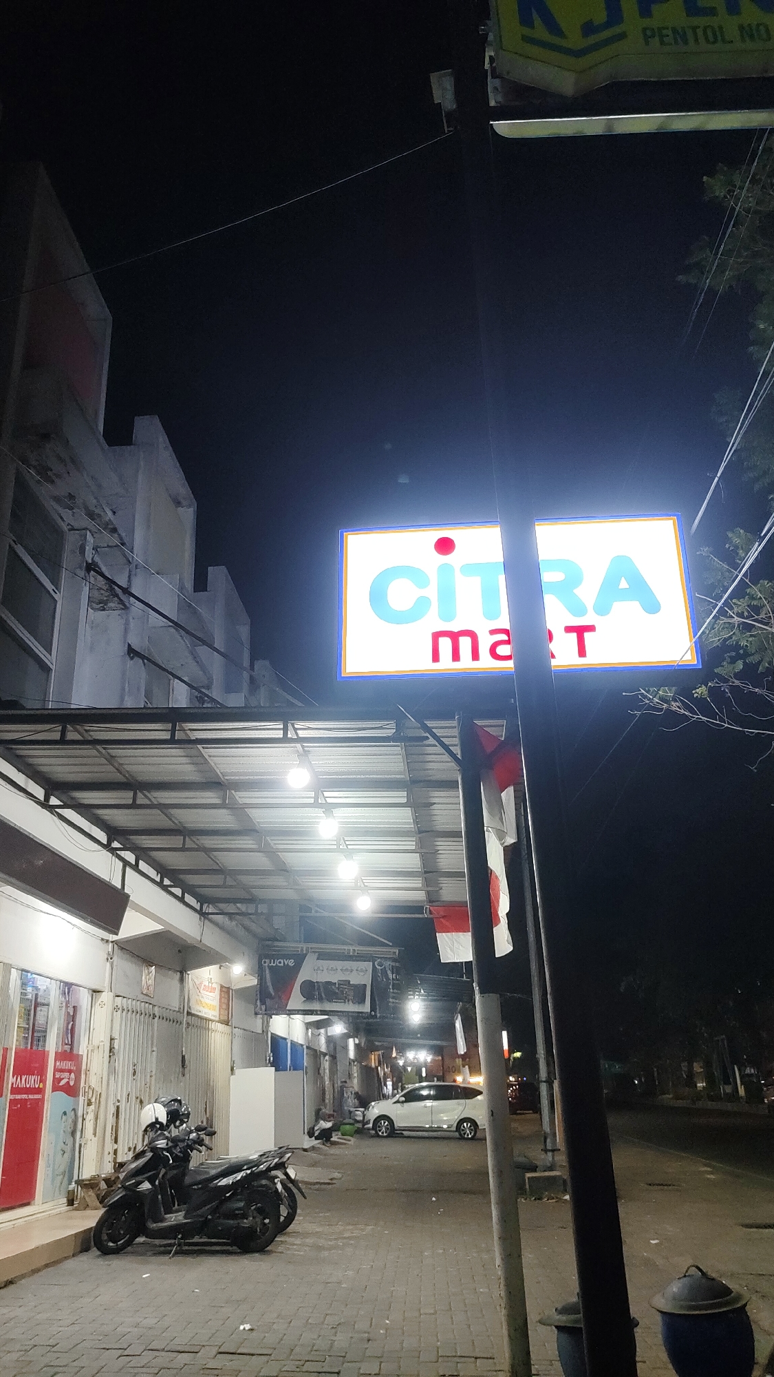 This is a nighttime exterior shot of a commercial establishment in Malang, Indonesia. A brightly lit sign with the words "CITRA mart" in blue and red lettering is prominently displayed. The establishment appears to be a store, with a covered walkway extending along its front, illuminated by several hanging lights. Parked on the sidewalk in front of the store is a black motorcycle with two helmets. A white car is also visible on the street. Indonesian flags are displayed near the sign, suggesting a local business or a patriotic display. The overall atmosphere is that of a typical urban commercial street scene at night.