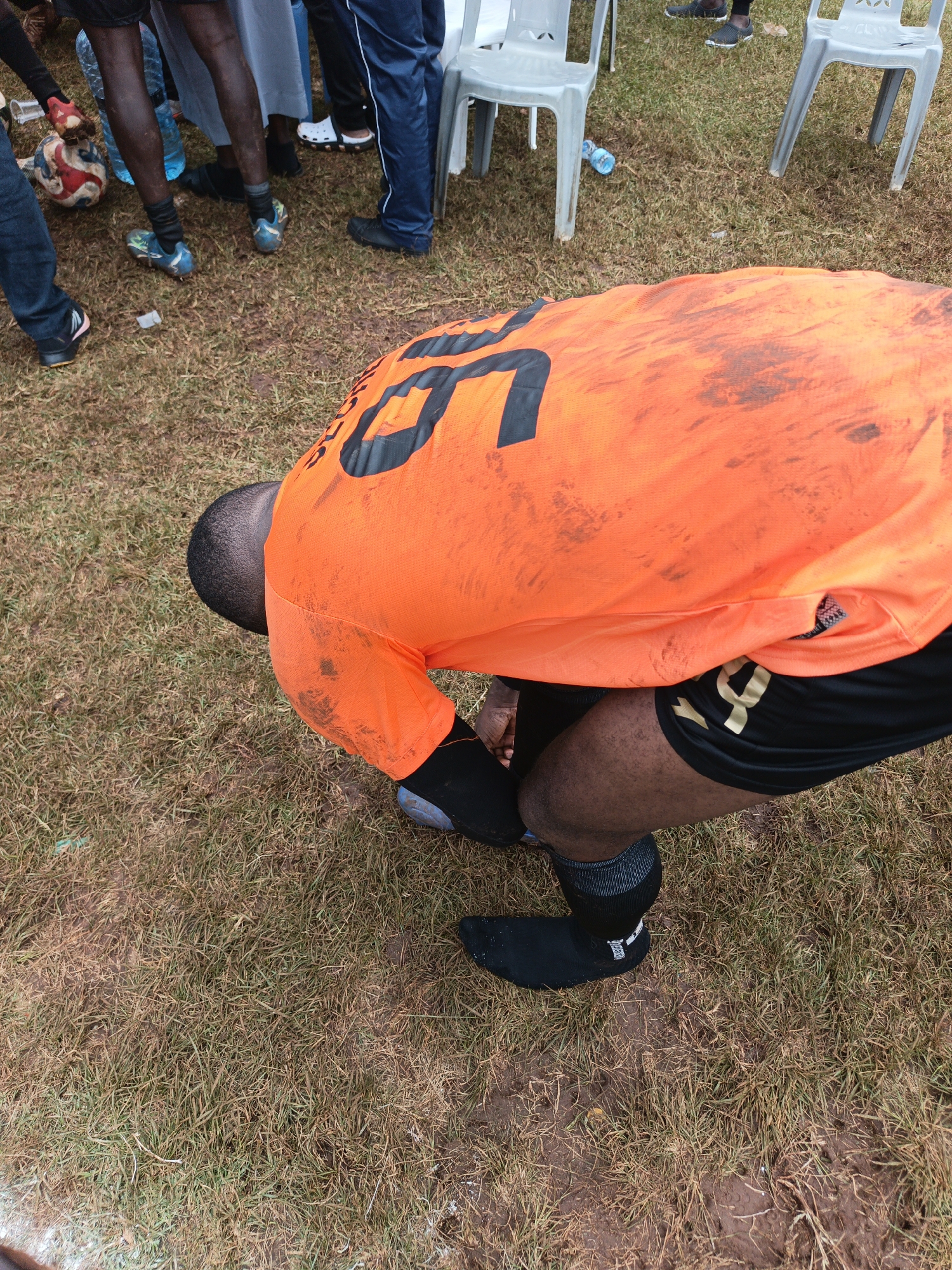 A man in an orange and black soccer uniform kneels on a grassy field, tying his shoe. The uniform is soiled with mud. Other people stand in the background near several white plastic chairs. The location appears to be Nairobi, Kenya, based on the environment and the context of a soccer game.