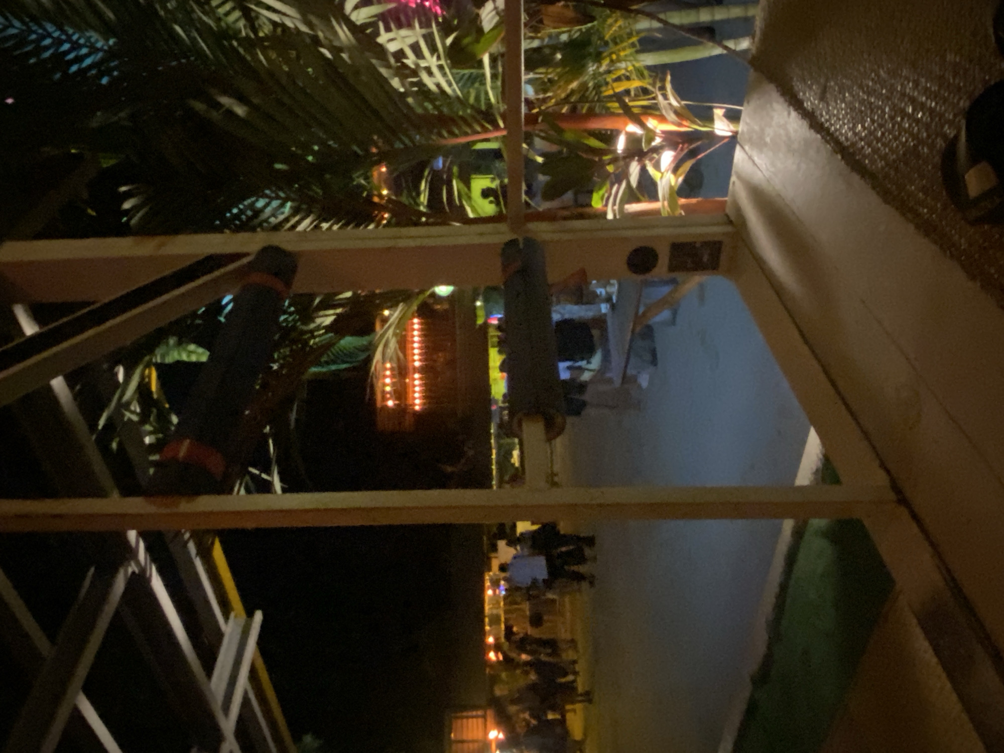 A sideways, nighttime, outdoor view taken from a bar or restaurant in Baan Hin Kong, Thailand, shows an outdoor area with people sitting at tables, with dim yellow lights illuminating the scene. Tables are occupied, suggesting social interaction. The view is framed by wooden beams and structures, with tropical vegetation and palm trees visible in the upper portion of the image.