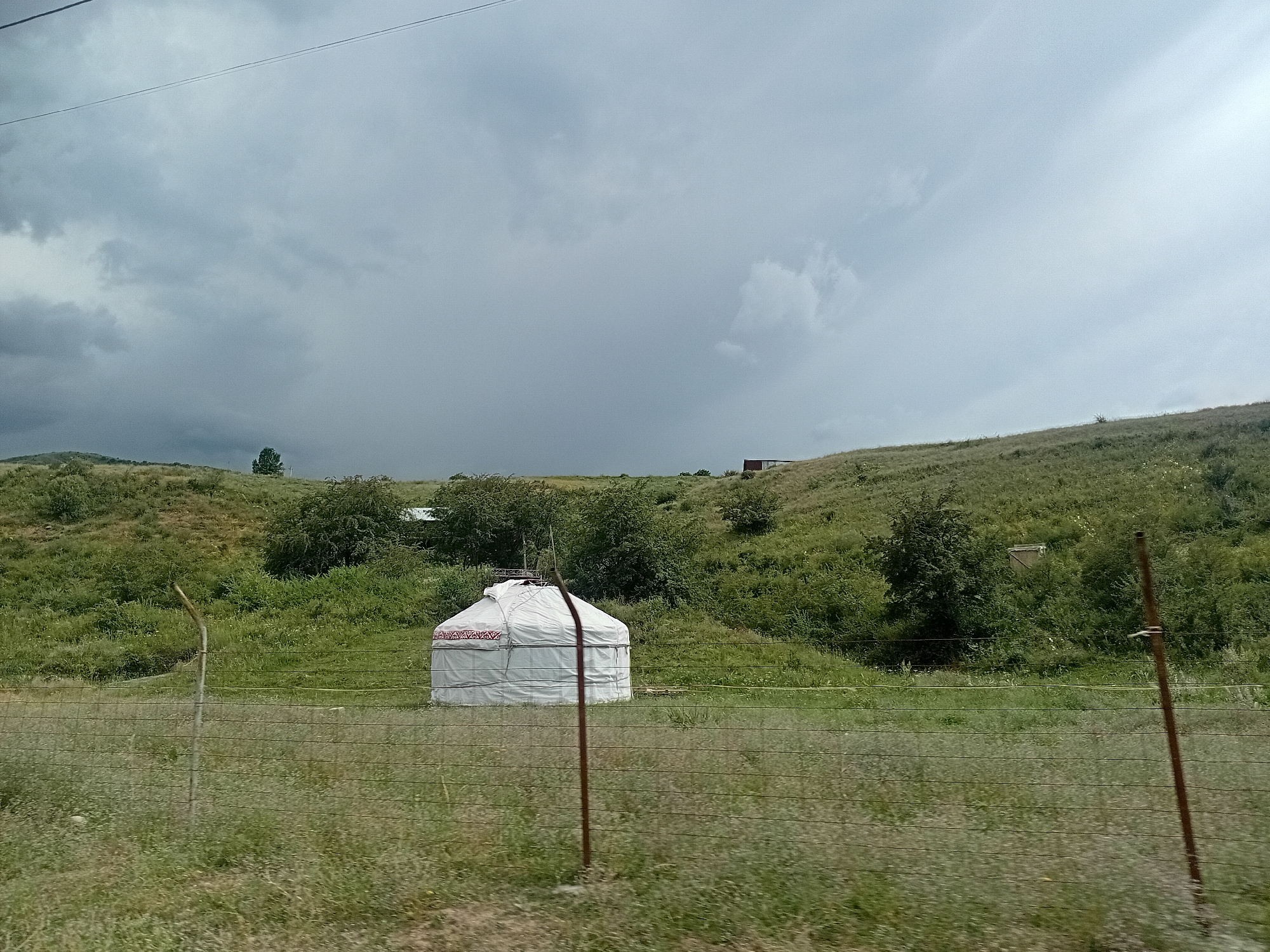 A wide shot captures a yurt, a traditional circular dwelling, situated in a grassy, rolling landscape under a heavily overcast sky. The yurt is predominantly white with a decorative red and white patterned band around its lower section. It is positioned in the middle ground, behind a wire fence that runs horizontally across the foreground. The fence is supported by metal posts. The terrain surrounding the yurt is uneven and covered in green grass and scattered bushes. In the distance, on the far right, a small, dark-colored structure is visible atop a hill. The sky is filled with dark, grey clouds, suggesting an impending storm or recent rain. A single, bare-branched tree stands on a distant hill to the left. The scene appears to be in a rural or semi-rural area, likely the region of Көксаяқ ауылдық округі, Kazakhstan, characterized by its open terrain and traditional dwellings. There are no people visible in the image.
