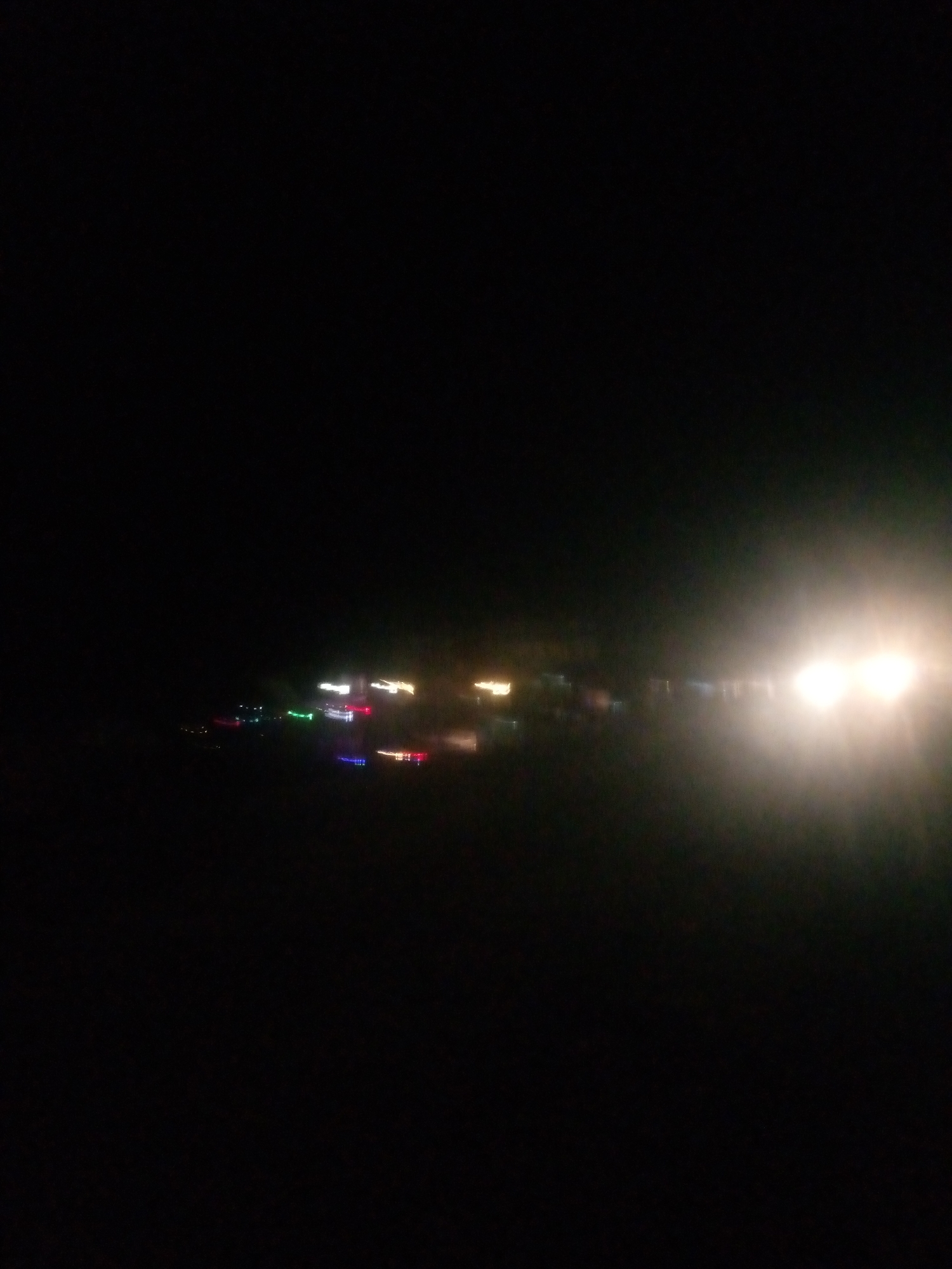 The image is a dark, out-of-focus photograph taken at night, likely from a moving vehicle. It captures a blur of lights, suggesting a street or highway scene. There are streaks of white, red, green, and blue lights, which are typical of vehicle headlights and taillights, as well as streetlights. In the far right of the image, there is a bright, diffused white light that could be from an oncoming vehicle's headlights or a large stationary light source. The overall impression is of a busy urban environment at night, with the motion blur indicating a degree of speed. The location in Jalingo, Nigeria, is not visually identifiable from the image itself, as there are no distinctive landmarks or features. The scene depicts a typical nocturnal urban landscape characterized by artificial illumination and the movement of vehicles.