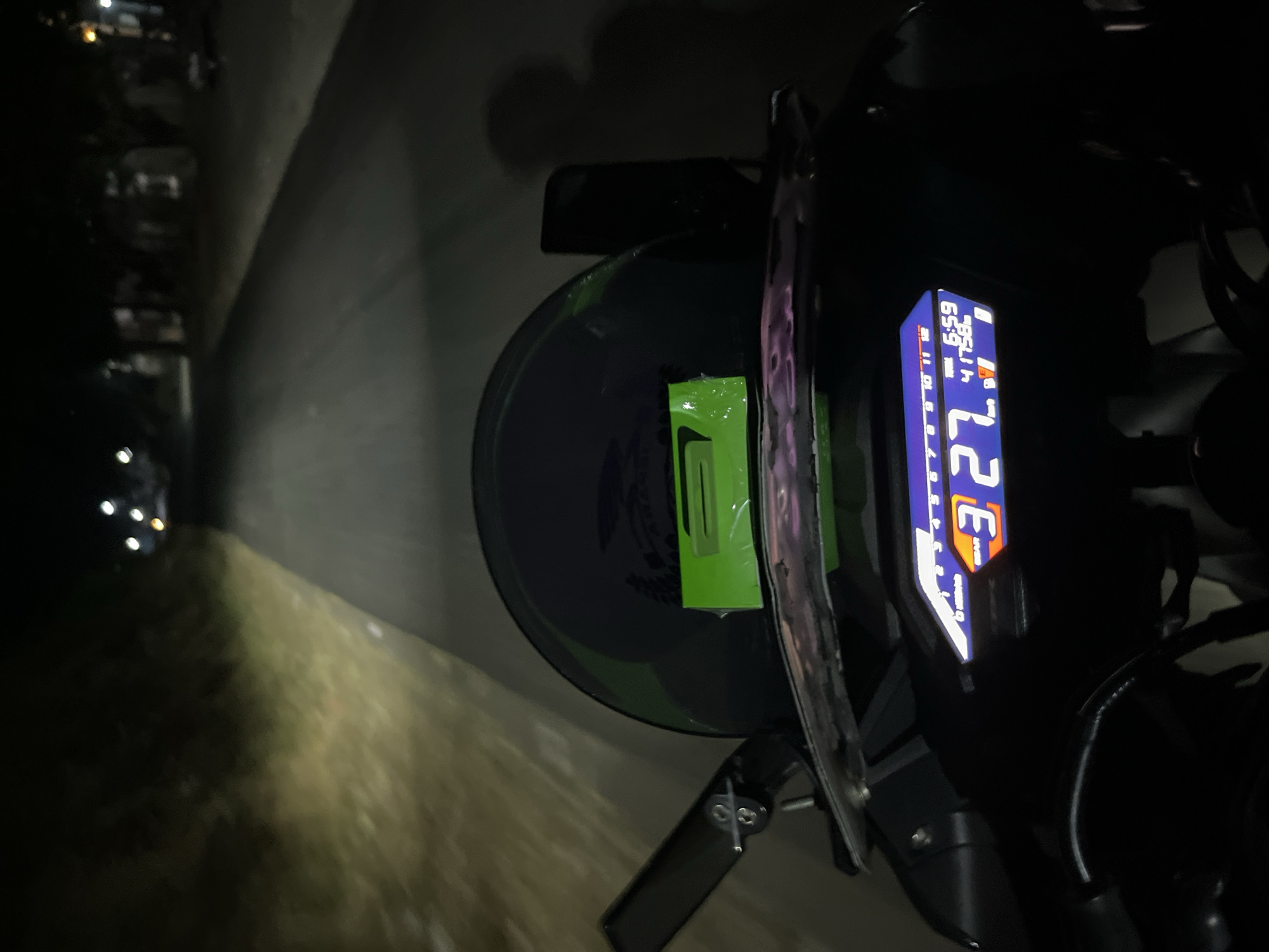 The image is a first-person perspective shot from a motorcycle at night, likely in Karangrandu, Indonesia, judging by the accompanying metadata. The motorcycle is on a paved road, illuminated by its headlight which casts a yellow-white glow on the left side of the road, highlighting sparse vegetation. The right side of the road is darker with indistinct structures and trees visible in the distance.
The motorcycle's dashboard is prominently displayed in the foreground, showing a digital speedometer reading "27" and "km/h" indicated by the "E" symbol. The time is displayed as "6:59", and an odometer reading of "41758" is also visible. A gear indicator shows "3". A green rectangular sticker is affixed to the helmet, which is positioned on the motorcycle. A dark, elongated object, possibly a phone, is mounted to the right of the helmet. A faint shadow of what appears to be a person is visible on the right side of the frame, projected onto a dark background. The overall scene suggests a night ride through a semi-urban or rural area.