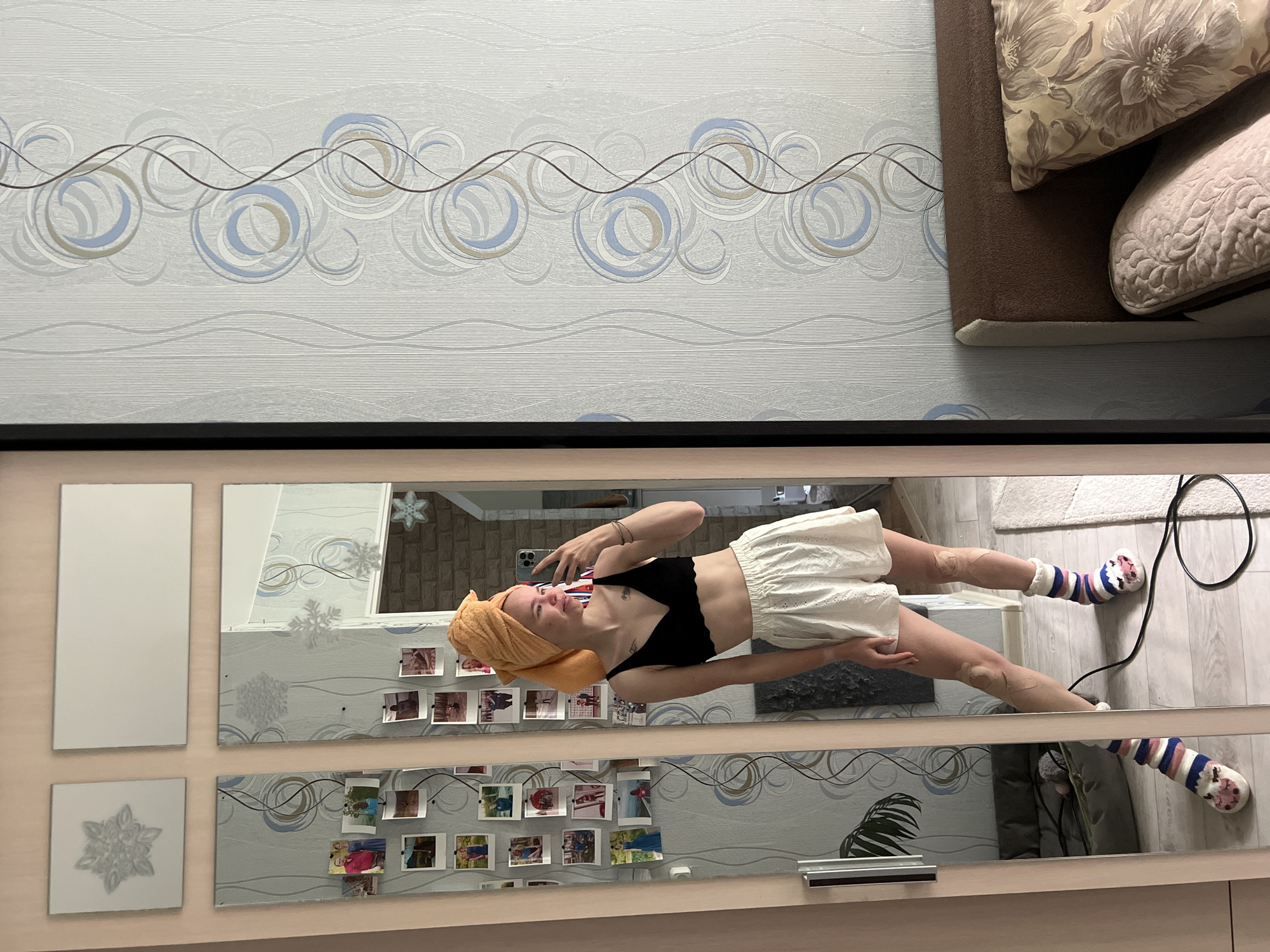 This media file presents a casual mirror selfie taken indoors in Chaikovsky, Russia. The original image is rotated, showing a young woman, likely in a bedroom or living space, reflected in a large mirror.
The primary subject is a light-skinned woman, standing upright in her reflection. Her hair is wrapped in an orange towel, suggesting she might have just showered. She is wearing a black scalloped bralette and high-waisted white shorts with eyelet details. Her feet are adorned with colorful striped socks featuring a cartoon-like character design. She holds a smartphone, appearing to be an iPhone, in her right hand, positioned to capture her reflection. Patches, possibly kinesiology tape, are visible on her legs. Her expression is neutral, with a slight upturn of her lips.
The mirror itself is set in a light wooden frame and comprises multiple panels. Several small, printed photographs, reminiscent of instant camera prints, are taped onto the mirror's surface, creating a personalized display. Decorative white snowflake cutouts are also attached to the mirror, hinting at a winter theme or decor.
The background, visible both directly and reflected in the mirror, features light blue and white wallpaper with a wavy pattern adorned with swirling circular designs in shades of blue, beige, and brown. In the upper right corner, a section of a dark brown couch or bed with two patterned cushions (one floral, one textured) is visible. The floor reflected in the lower part of the mirror is light-colored wood, partially covered by a white textured rug. A black electrical cable lies on the floor near the woman's feet. Beyond the mirror, a wall with a brick-like pattern and a window frame are visible in the reflection.
No specific time of day is discernible, but the lighting suggests daytime. There is no visible text in the image. The overall scene conveys a relaxed, domestic atmosphere.
