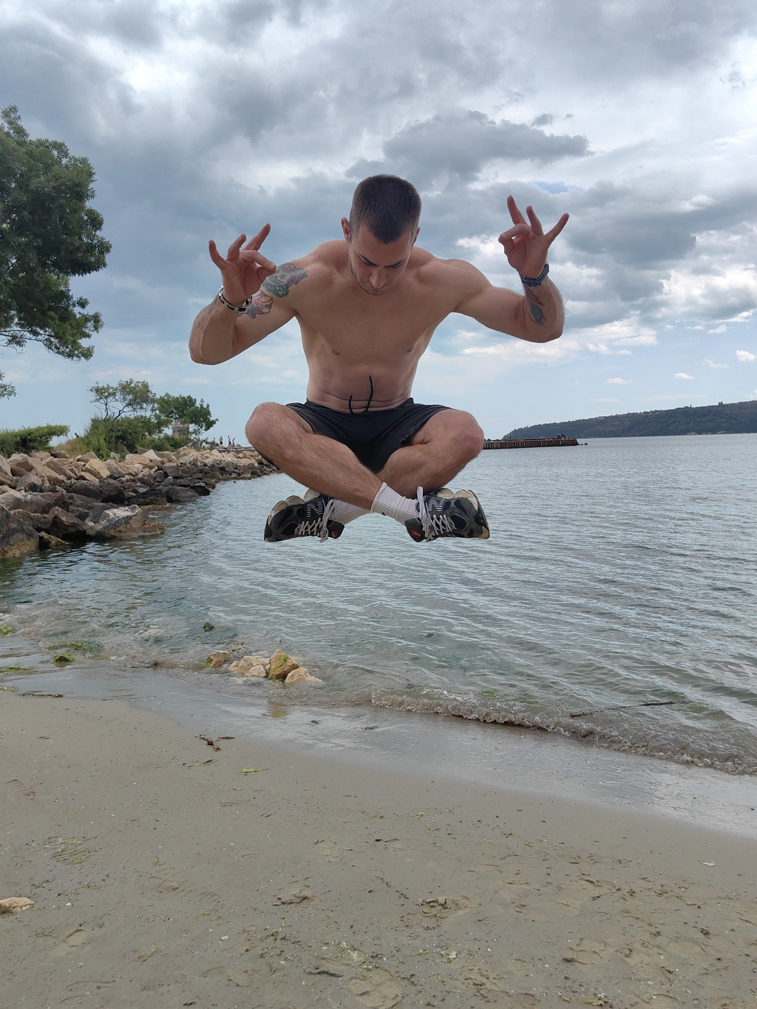 A shirtless man in black shorts, sneakers, and white socks is suspended in mid-air in a cross-legged position, with his hands raised, making a hand gesture on a beach in Varna, Bulgaria. He has tattoos on his arms. The beach is sandy with some rocks and vegetation near the water's edge. The water is slightly choppy with waves. In the background, there is a body of water, possibly the Black Sea, and a distant shoreline with some structures visible. The sky is overcast with clouds.