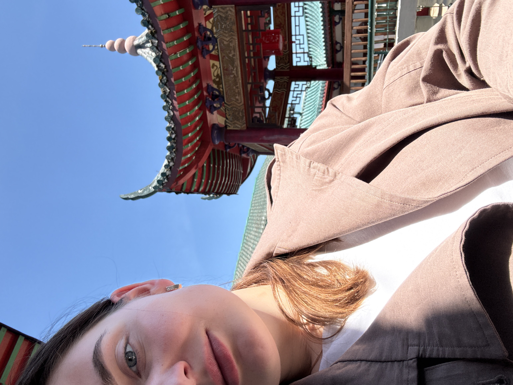 A person is captured in a low-angle selfie, with part of her face, brown hair, and shoulder visible on the left and lower right of the frame. She has fair skin, wears a small ear piercing, and is dressed in a light brown or taupe jacket over a white top. The image is taken outdoors on a bright, sunny day, indicated by the clear blue sky and strong daylight.
In the background, dominating the upper and right portions of the frame, is a vibrant building with distinct East Asian architectural features. The structure showcases a multi-tiered roof adorned with red and green tiles or decorative elements, intricate wooden beams, and latticework. A slender finial or spire rises from the top of the roof against the sky.
The scene suggests a moment of leisure, possibly a tourist or visitor taking a picture with the decorative architecture. Given the provided location context of Hainburg an der Donau, Austria, this building is likely an East Asian-style temple or pagoda situated within that area, such as the Hainburg Pagoda or Buddhist Center. No visible text is present in the image.