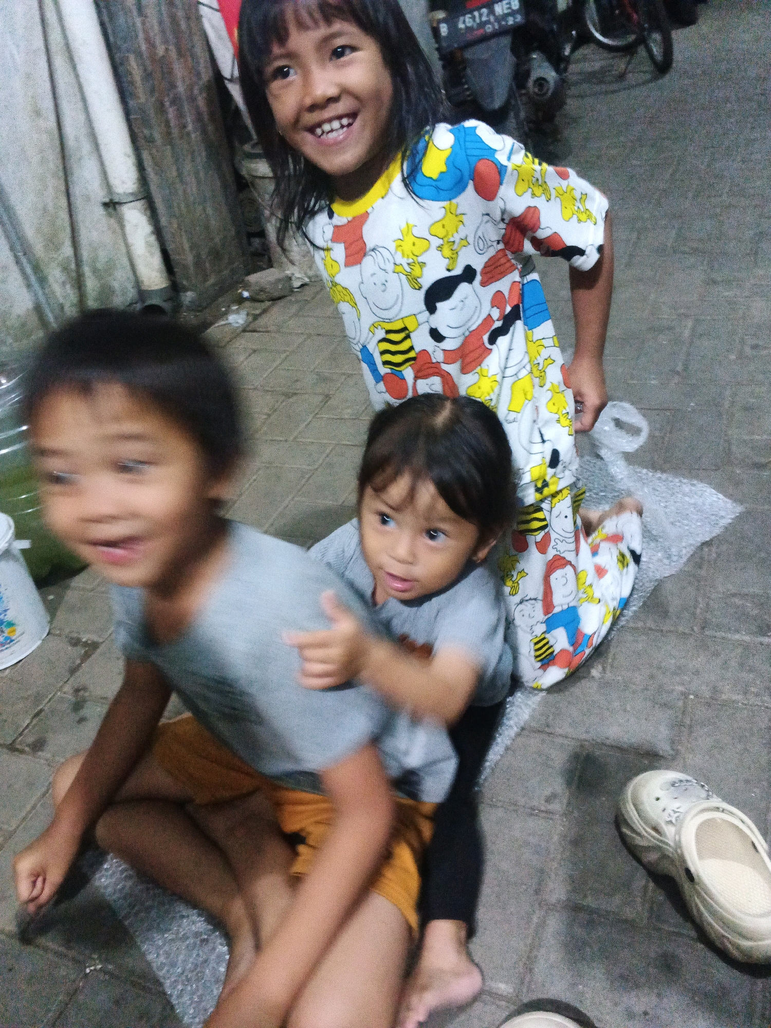 In the city of Kadu, Indonesia, two young children are captured in a close-up, outdoor scene. The child in the foreground, on the left, is a young girl with short, dark hair and a bright smile, wearing a gray t-shirt and orange shorts. She is seated, and her body is slightly blurred due to motion. To her right, another young child, also with dark hair, is seated, leaning against the first child. This child is wearing a gray t-shirt and black leggings. They are looking to the left.
The elder child, standing behind the seated children and slightly to the right, is a young girl with long, dark hair. She is wearing a white pajama set adorned with a cartoon pattern featuring Snoopy and Woodstock. She is smiling broadly and looking towards the camera.
The environment appears to be a paved outdoor area, possibly a street or alleyway, with a concrete or tiled ground. In the background, a motorcycle is partially visible, along with some corrugated metal sheeting and a wall. There is also a piece of bubble wrap on the ground. A light-colored clog is visible on the right side of the frame. The lighting suggests it might be evening or a dimly lit area.