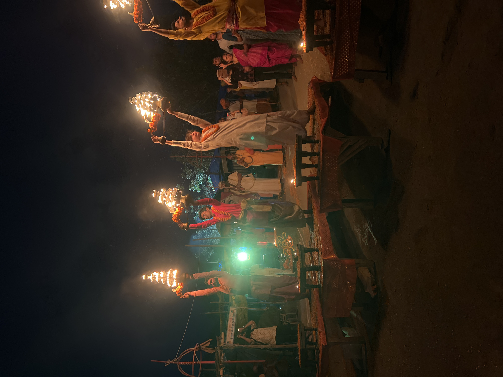 The image captures a night scene in Vrindavan, India, showcasing a Hindu ritualistic ceremony. Four men stand on elevated platforms, each holding a multi-tiered lamp with flames. Each platform has an ornate, red-colored covering. They are adorned with garlands of orange flowers. Below, a group of people are gathered, some holding cameras, while others appear to be observing the ceremony. The background is a mix of dark sky and indistinct structures. The lighting is predominantly provided by the flames from the lamps, casting a glow on the participants and the immediate surroundings.
