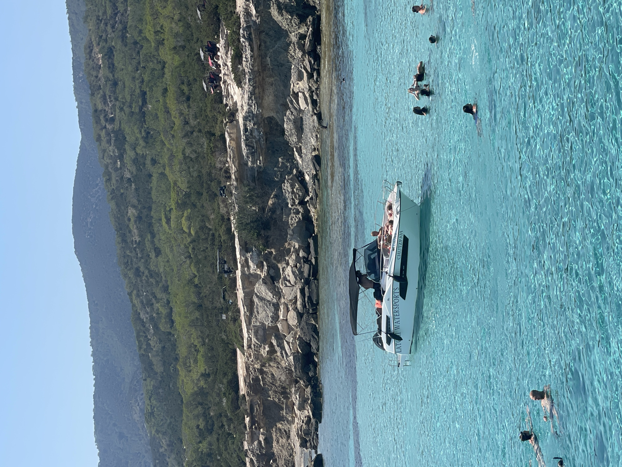 A bright, sunny day at a tropical coastal location, likely in Neo Chorio, Cyprus, shows a calm, turquoise sea filled with people swimming and a white boat. The boat, labeled "LATO WATERSPORTS" and "CHUTZPAH," has a dark canopy and several people on board, some sunbathing and others standing. In the background, rugged, rocky cliffs rise up, covered with lush green vegetation and trees. Atop the cliffs, several all-terrain vehicles (ATVs) are parked, suggesting a nearby recreational area. The water is crystal clear, allowing visibility of the seabed in some areas and the swimmers beneath the surface. The sky is a clear, pale blue.
