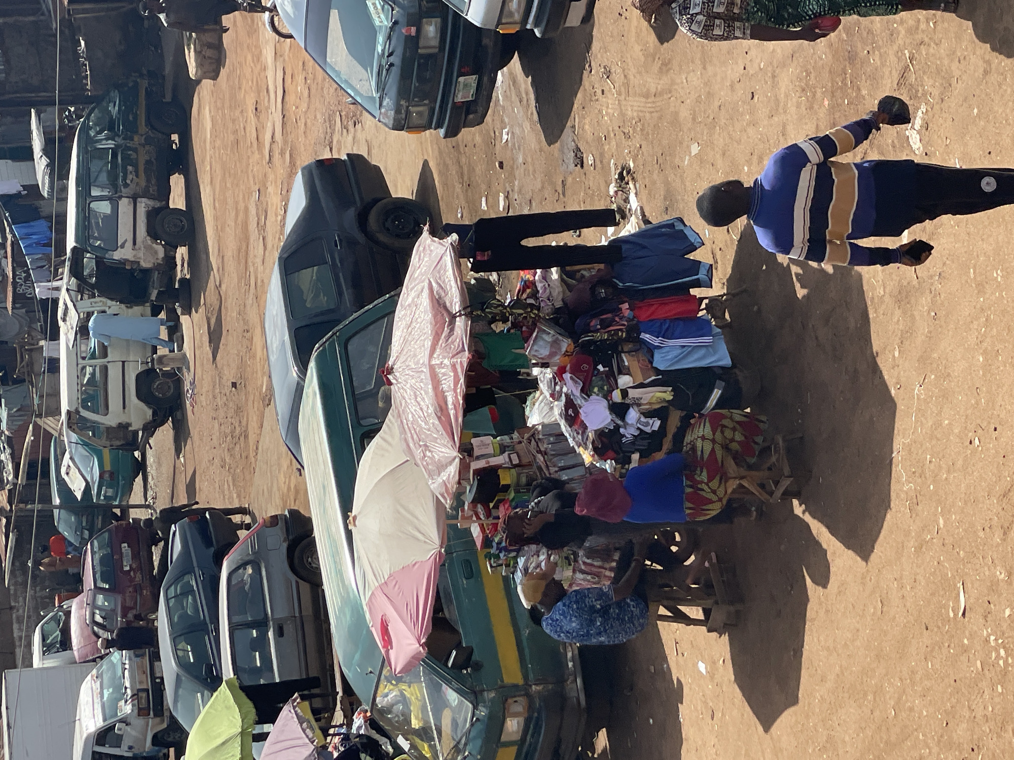 An overhead shot captures a bustling outdoor market in a city. The ground is a dusty, reddish-brown dirt, littered with small pieces of debris. Several vehicles, including vans and cars, are parked or driving through the scene, some appearing worn and aged. A prominent green and yellow van with an open back sits in the center, displaying various goods under two colorful umbrellas. People are seen milling around, some attending to the market stalls, while others walk through the area. One person, wearing a blue and white striped sweater, walks away from the camera, holding a dark object in their hand. Another person in a blue top and a patterned skirt is seated at a stall. The overall impression is of a busy, vibrant marketplace with a focus on commerce and daily life.
