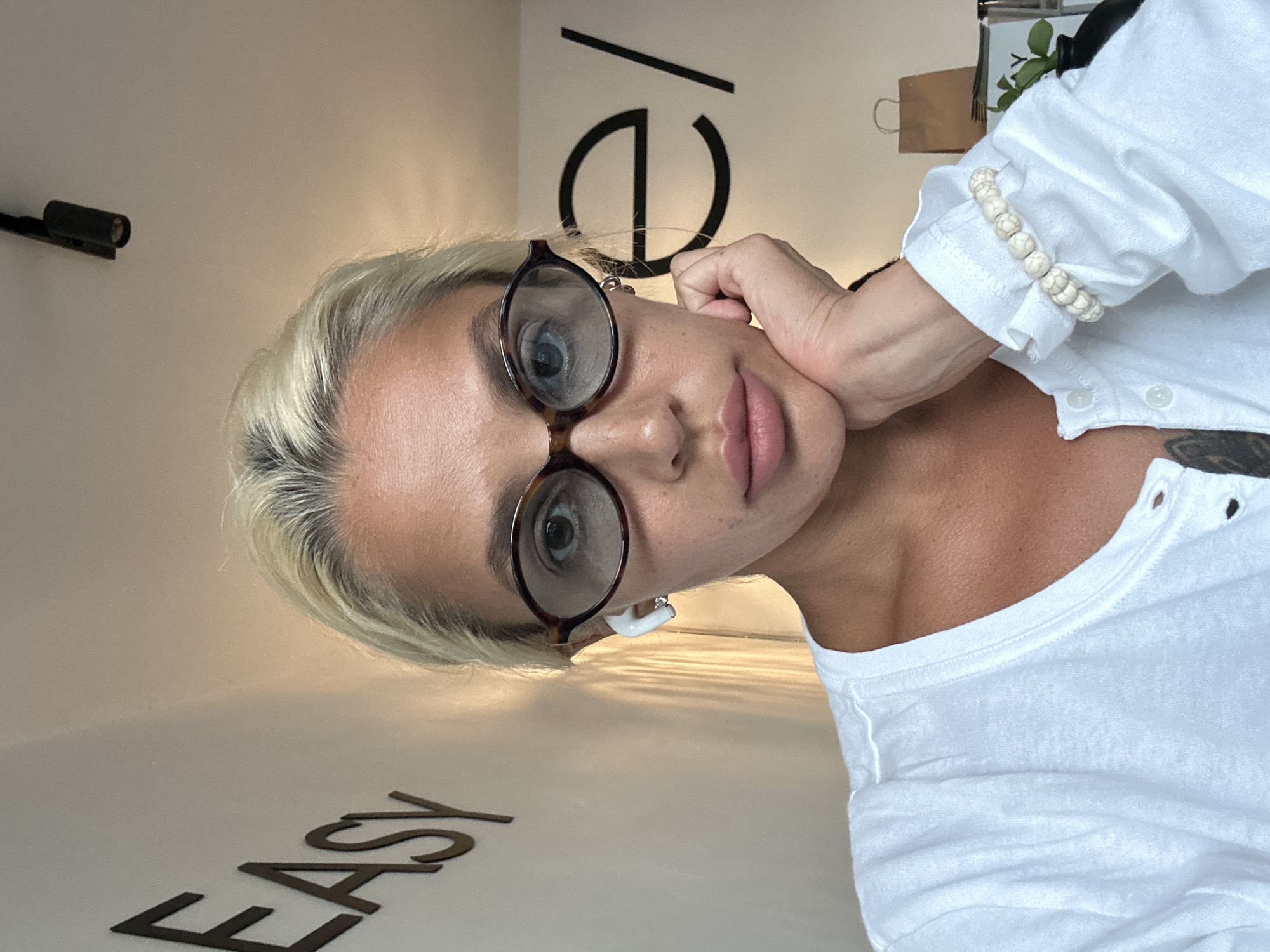 A woman with blonde hair and glasses is shown in a selfie. She is wearing a white shirt and has a bracelet on her left wrist. She is also wearing white earbuds. Behind her, the word "EASY" is visible on the wall in large letters, and the letters "e/" are also visible. The scene appears to be indoors, possibly an office or home environment. There is a paper bag and a plant in the background. The lighting suggests it is daytime. No specific location in Kuta Selatan, Indonesia can be determined from the visual information provided.