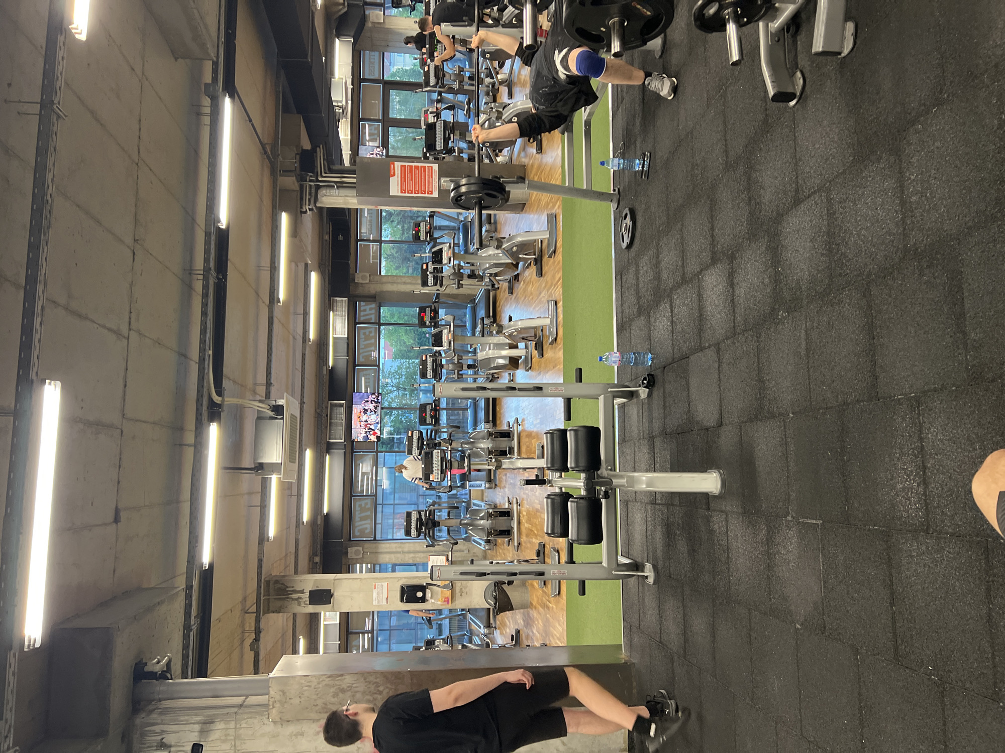 The image shows the interior of a gym. Several people are exercising on various weight machines. There are several rows of machines throughout the gym. Concrete walls are visible on the left side. Windows line the back wall. There are rows of fluorescent lights on the ceiling. The city is Sofia, Bulgaria.