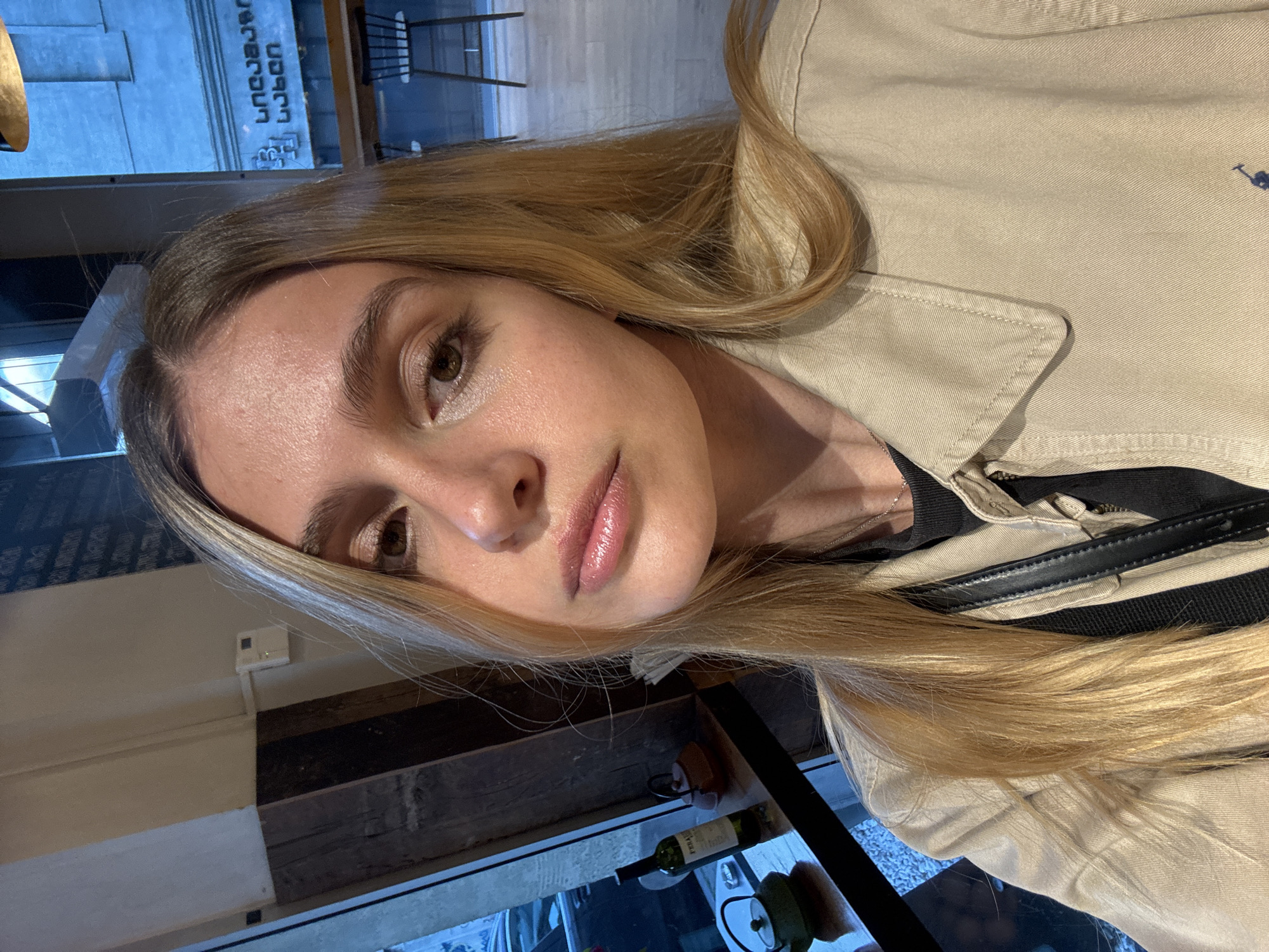 A young woman with long, blonde hair is posing for a selfie inside a restaurant or cafe. She's wearing a tan jacket, a black top, and a crossbody bag with a black strap. A small portion of a bottle of wine and other glassware are visible on the table. Outside the window, there's a building with Georgian text. The location appears to be Tbilisi, Georgia.