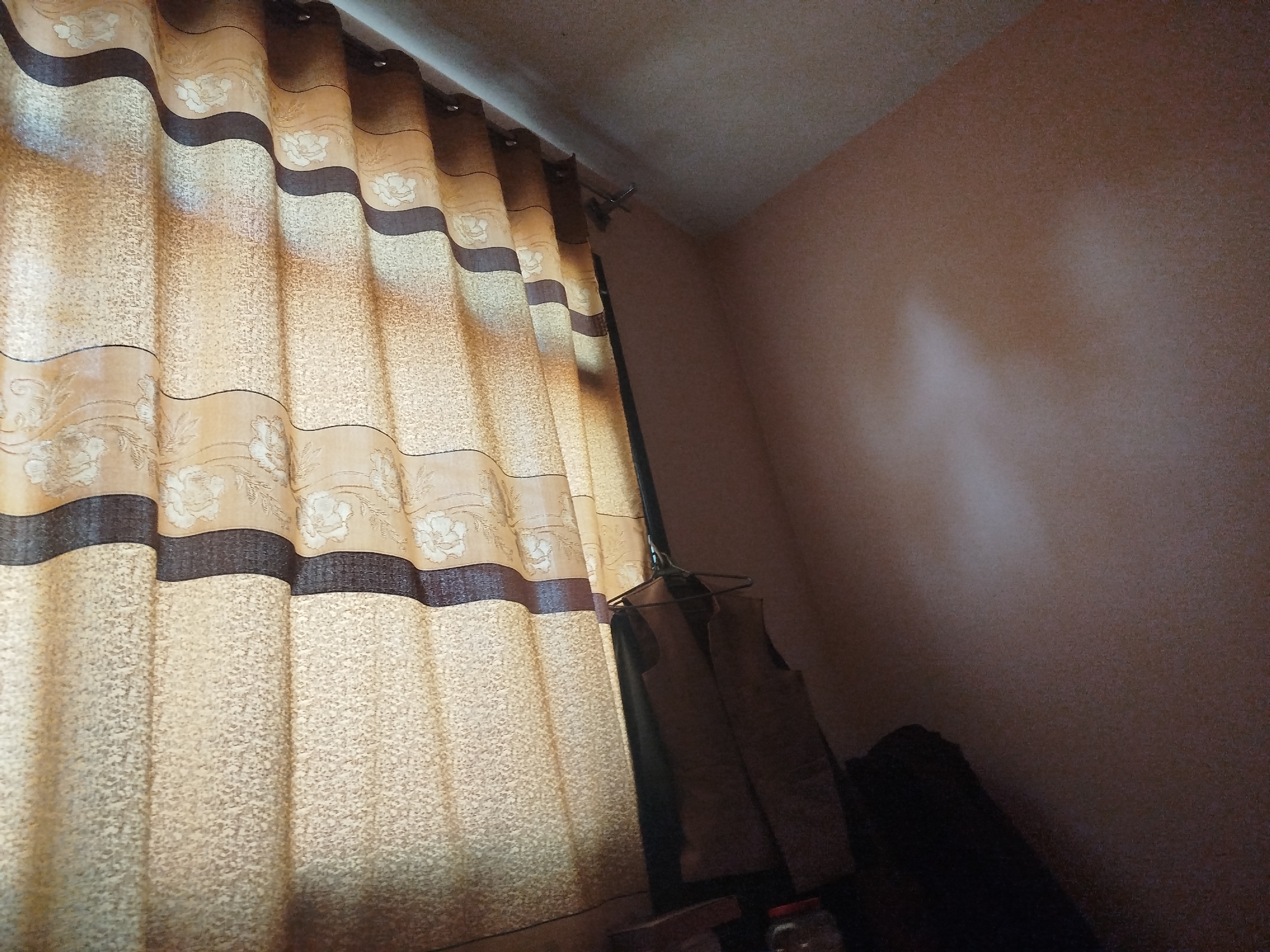 In the city of Lassan, Pakistan, an indoor scene is depicted, focusing on a window with a patterned curtain. The curtain, primarily beige with dark brown horizontal stripes and floral motifs, is drawn to the left, revealing a portion of the wall and ceiling. A dark garment hangs on a hanger from a rod in front of the curtain. The wall is painted a light brown or peach color, and there is a visible shadow or light reflection on it. The ceiling is also visible and appears to be a light color. The overall lighting suggests an indoor environment, possibly a home.