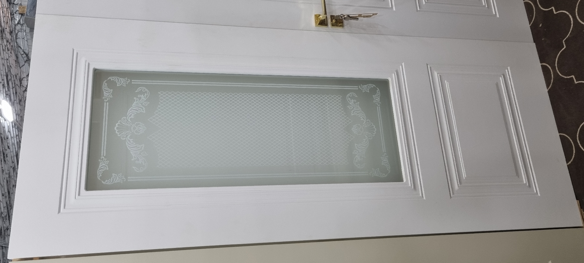 The image shows a close-up of a white interior door with decorative frosted glass. The glass has an ornate pattern that includes floral motifs and a cross-hatch design. The door is paneled and has a gold-colored door handle with keys attached. The flooring visible is a dark carpet with a light-colored quatrefoil pattern. The background appears to be a textured wall. There are no people or specific landmarks visible that would indicate the city of Bokhtar, Tajikistan.