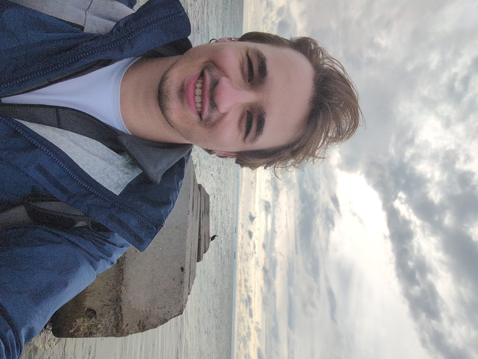A young man with short, messy brown hair and a light beard smiles at the camera. He is wearing a dark blue jacket over a white t-shirt and has hoop earrings in both ears. He is standing in front of a grey concrete structure extending into the sea. In the background, the sea is calm under a cloudy sky, with several ships visible on the horizon. A bird is perched on the concrete structure in the middle ground. The location is likely Odesa, Ukraine, given the architectural style of the concrete structure and the presence of the Black Sea.