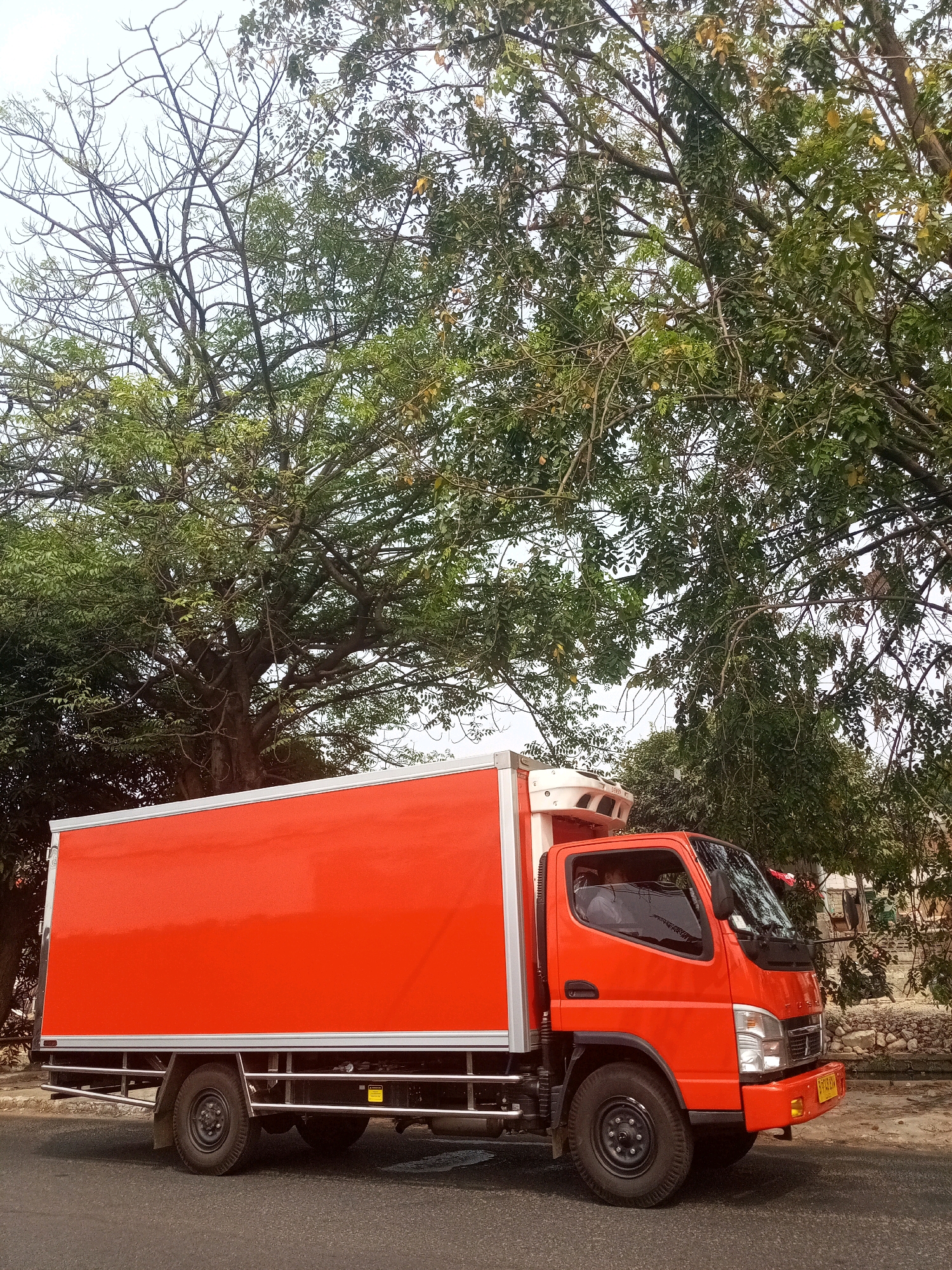 An orange refrigerated truck is parked on the side of a road in Bekasi, Indonesia. The truck has a white roof and silver trim along the side of the cargo box. The driver's side door is visible, and a person can be seen inside the cab. The truck is positioned in front of lush green trees, some of which appear to be bare or have sparse foliage. The sky is overcast. The ground is paved with asphalt.