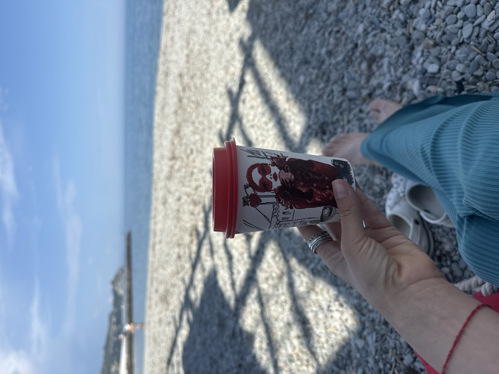The image shows a person holding a disposable coffee cup with a red lid in their right hand, adorned with multiple silver rings, on a pebble beach. The cup's design appears to be black and white with red accents. To the right, there is a bare foot, a white shoe, and a portion of turquoise clothing. In the background, the body of water extends to the horizon under a blue sky with light clouds. The scene suggests a relaxed setting, possibly on a shoreline. Location is Unknown, Unknown.