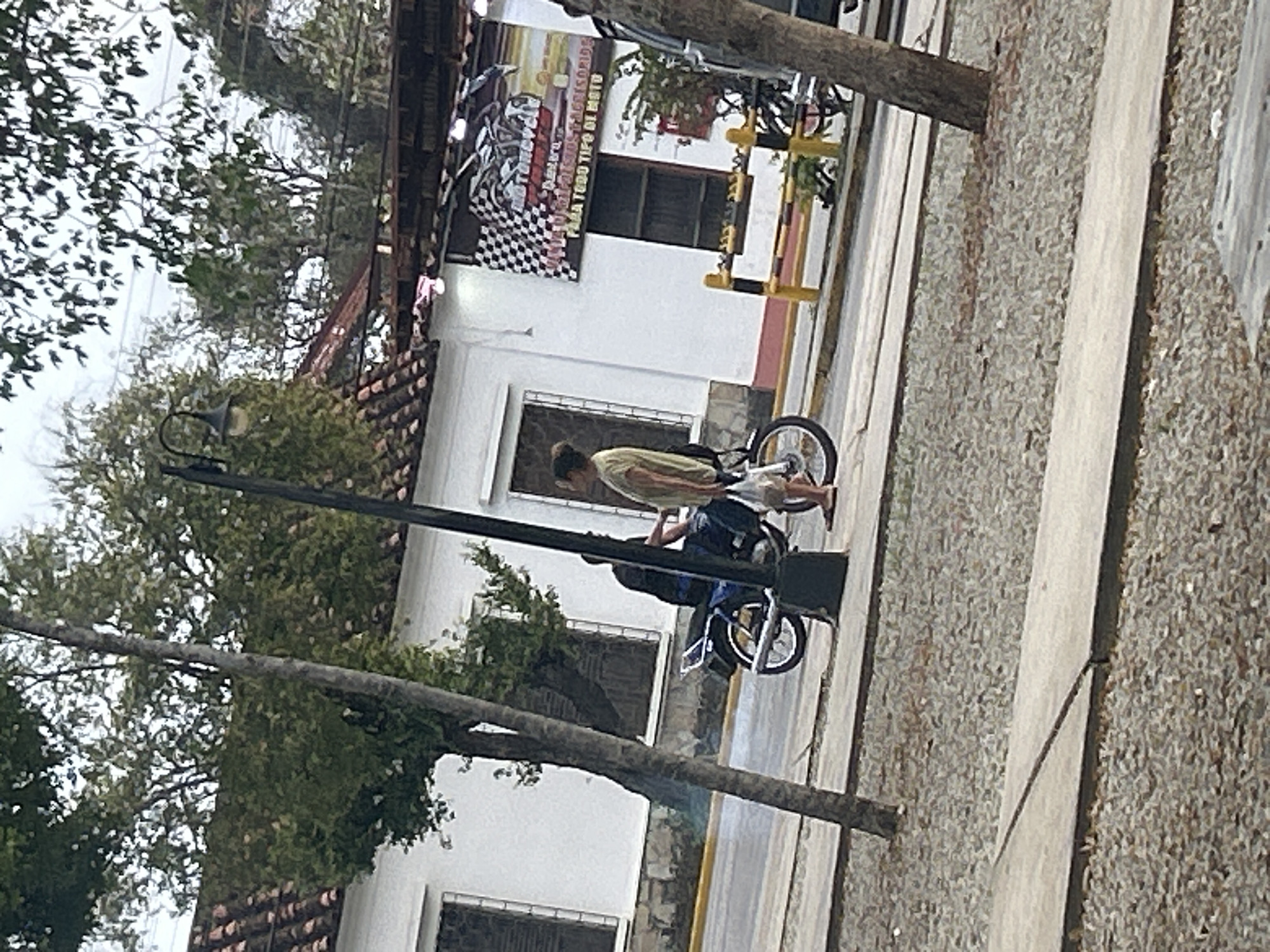 A motorcycle is parked on a street in Santa Ana, Venezuela. A person wearing a light-colored shirt and shorts stands next to the motorcycle, holding a plastic bag. Another person sits on the motorcycle. A building with a white facade and a red-tiled roof is visible behind them. A street lamp is positioned to the left of the scene. The street appears to be paved, with a sidewalk made of concrete slabs and gravel. Trees and foliage are present in the background. A sign with a checkered flag design and text in Spanish is mounted on the building. The overall scene depicts a street-side view in a town or city.