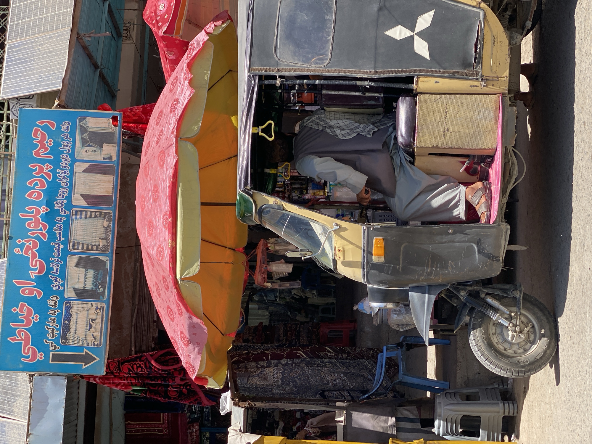 In Kandahar, Afghanistan, a street scene unfolds under a bright, sunny sky. A three-wheeled auto-rickshaw, predominantly black with yellow accents, is parked. The driver, wearing a grey vest over a light blue shirt and a patterned cap, is seated behind the wheel. His legs are clad in grey trousers, and he wears brown sandals. A checkered cloth is draped over his shoulder. The auto-rickshaw's interior is visible, revealing shelves stocked with various bottles and items. A red and yellow umbrella, offering shade, is positioned above the vehicle and extends over a market stall.
To the left of the auto-rickshaw, a market stall displays colorful textiles, including red blankets with intricate patterns. A blue plastic chair and a grey plastic stool are also present in front of the stall. Behind the auto-rickshaw, a blue sign with Arabic writing is prominently displayed, featuring an arrow pointing downwards and three smaller images of curtains. The overall impression is a busy marketplace, capturing a moment of everyday life in Kandahar.