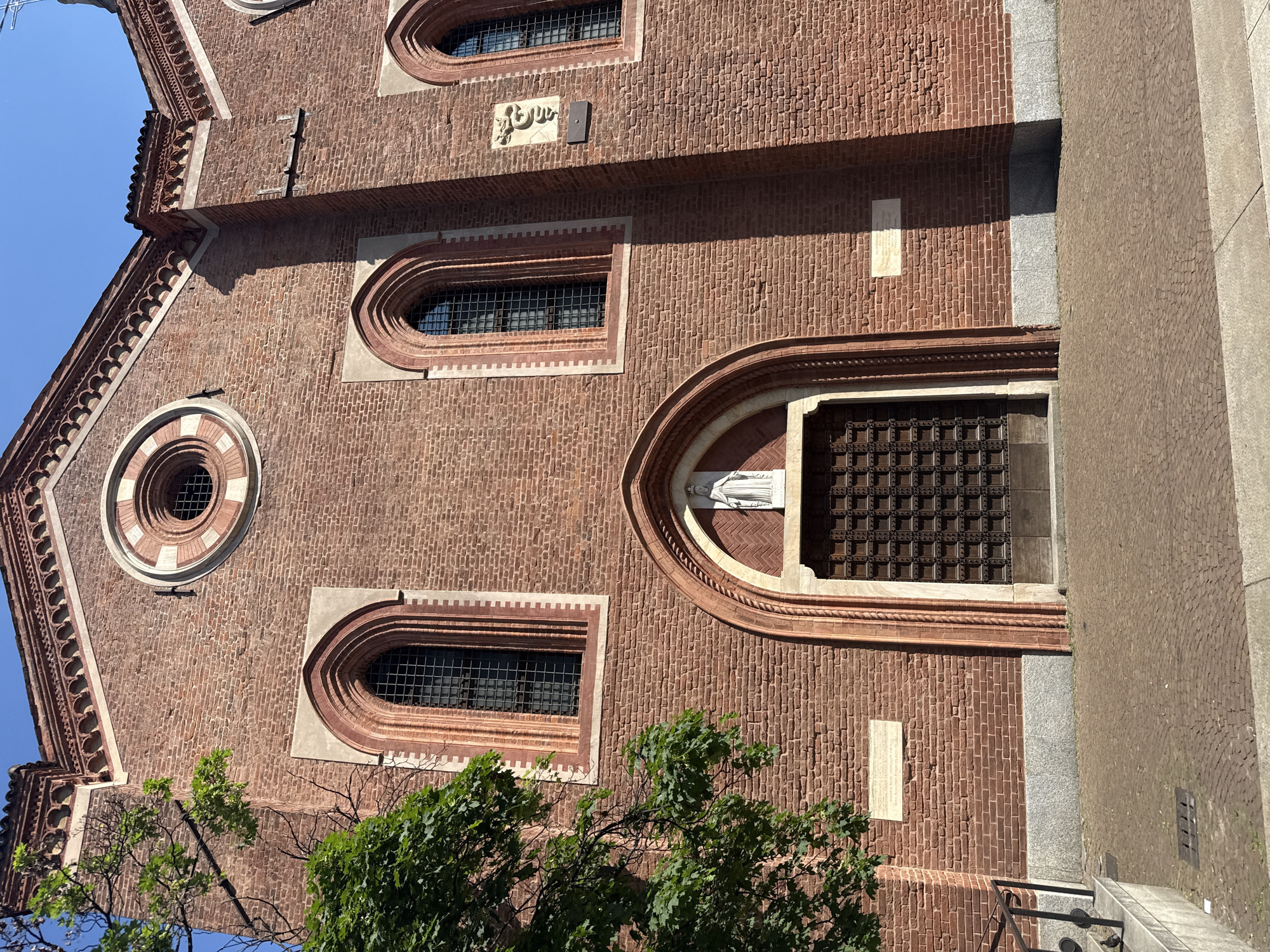 The image displays the brick facade of a historic building, likely a church or monastery, under clear blue skies in bright daylight. The structure is predominantly made of reddish-brown brick, featuring several arched windows with dark grilles and a prominent round oculus window, all framed by lighter-colored stone.
A notable architectural element is a large arched entrance on the right side, housing an ornate, dark wooden door with a square grid pattern. Above this entrance, within a decorative arched niche, stands a white statue of a robed figure, presumably a religious figure like the Virgin Mary.
On the brick wall above one of the arched windows, there is a distinct bas-relief carving of a serpent or dragon-like creature, often associated with the Visconti family symbol (Biscione) common in Milan. Adjacent to this carving is a small, dark rectangular plaque. Another light-colored rectangular plaque with faint, unreadable inscribed text is embedded in the brickwork to the left of the main entrance.
The building's base includes gray stone elements, with steps leading down to a patterned ground surface, possibly cobblestones or paving, on the right. Green foliage from trees partially obscures the lower left and central portions of the facade. Strong shadows are cast across the building, indicating a sunny day, likely late morning or early afternoon. No human activity is visible. The architectural style and potential Visconti symbol align with the given context of Milan, Italy.