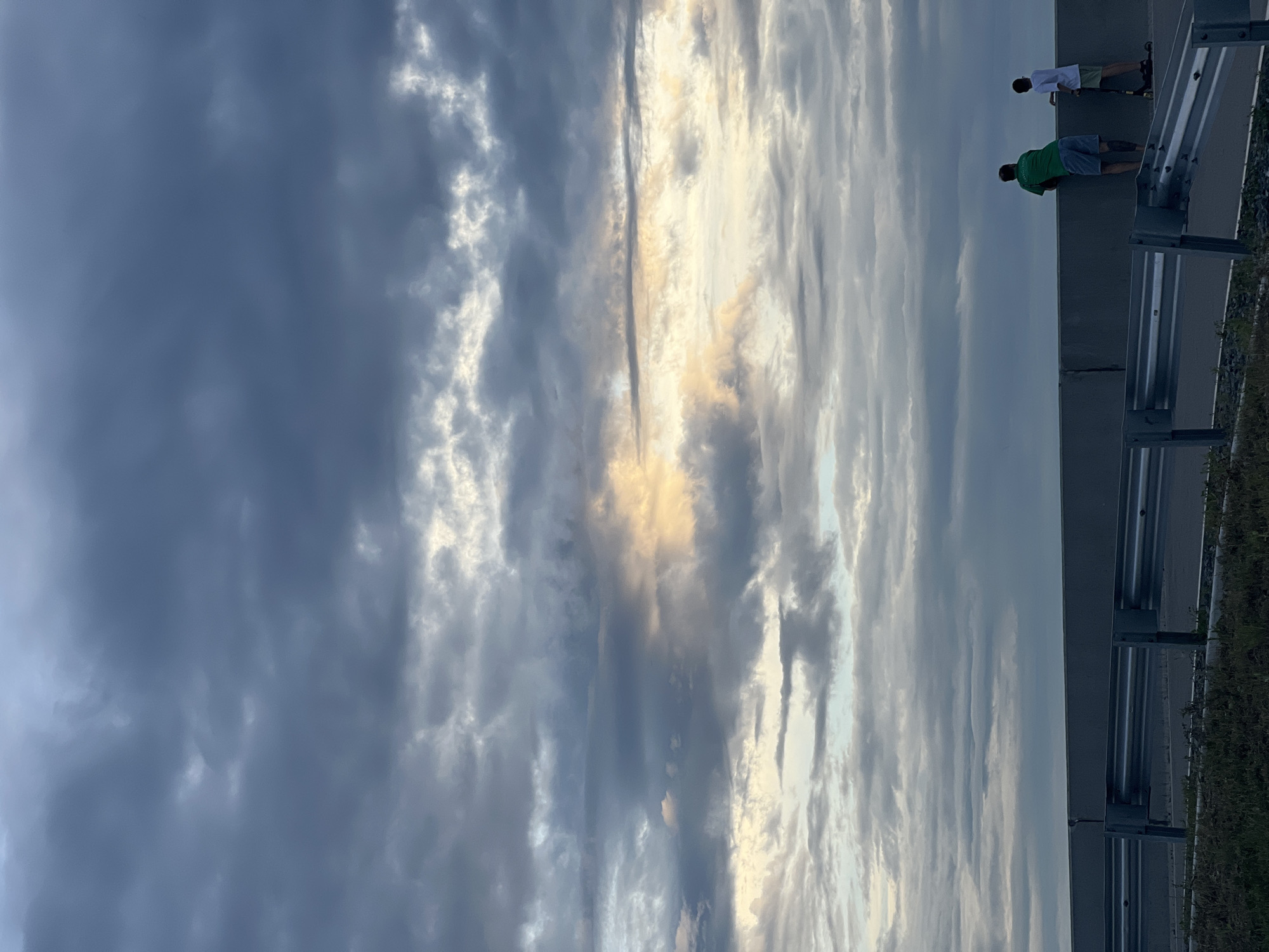 The image depicts a sky filled with dramatic, layered clouds in various shades of grey and white, with hints of yellow light breaking through. In the foreground, two individuals are standing on a raised platform or bridge, looking out at the sky. One person is wearing a green t-shirt and blue shorts, and the other is wearing a white t-shirt and light-colored shorts. The person in the white t-shirt is standing next to a scooter. A metal guardrail is visible in front of them, suggesting they are on a roadway or elevated walkway. The ground in front of the guardrail is covered in sparse grass. The scene appears to be set during twilight or an overcast day, given the lighting conditions. The location of Khabarovsk, Russia, is not visually represented in the image.