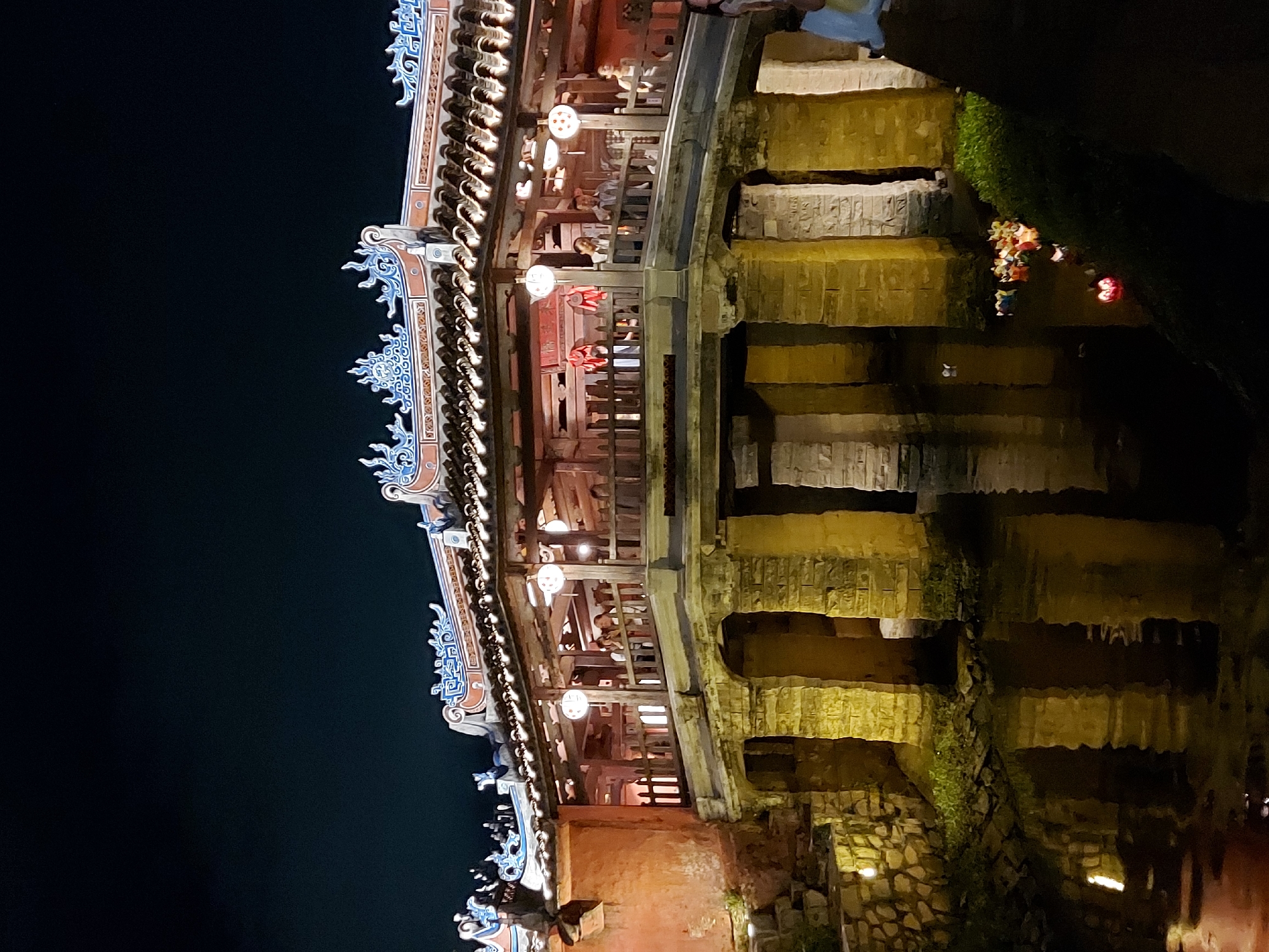 The image shows the Japanese Covered Bridge in Hoi An, Vietnam, at night. The bridge is illuminated by lights, highlighting its architectural details. The bridge has a tiled roof with ornate carvings and is supported by stone pillars over a dark body of water. Several paper lanterns hang from the ceiling of the covered walkway on the bridge, casting a warm glow. Inside the bridge, several people are visible, standing and looking around. In the water below, a cluster of floating lanterns creates a colorful display, with individual lanterns also visible floating separately. The surrounding area is dark, with the exception of some lighting on the stone walls and a patch of green grass on the right bank.