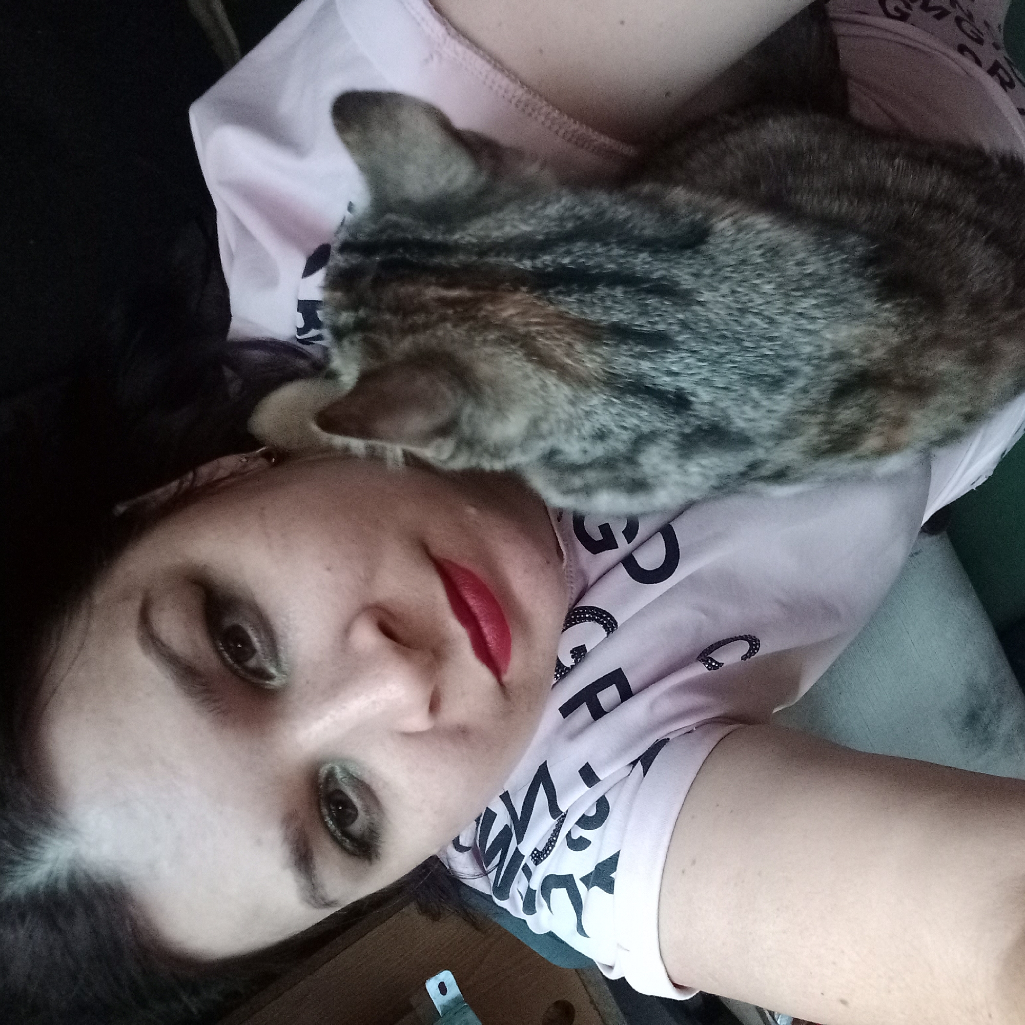 In an indoor setting, an individual lies on their back with a tabby cat resting on their chest. The individual is wearing a light pink t-shirt with black lettering. The cat's fur displays stripes of gray, black, and brown. The individual has dark hair and is wearing red lipstick and eye makeup. The background consists of a blurred surface, possibly furniture. The image is cropped to focus on the individual and the cat.