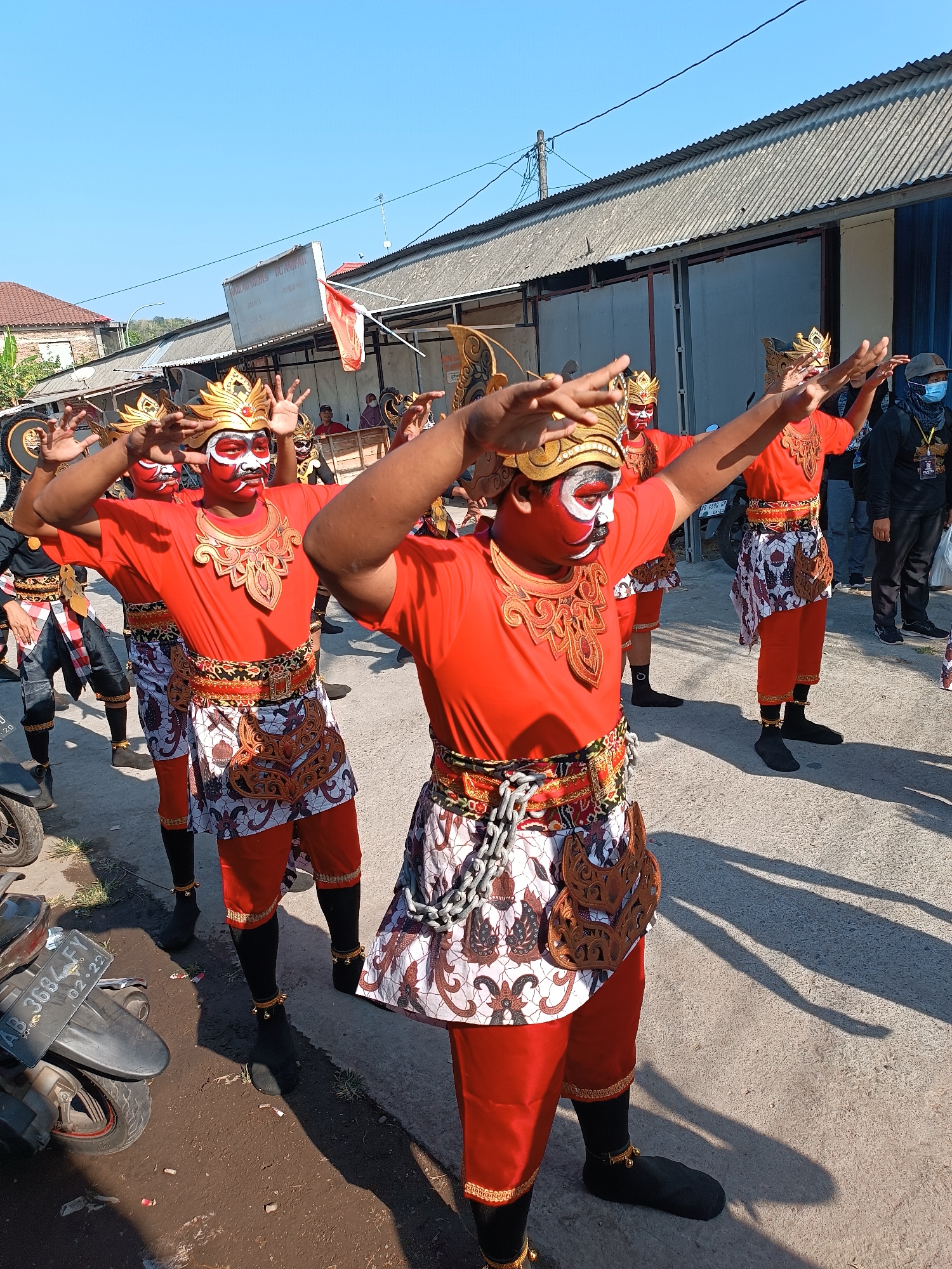 In Imogiri, Indonesia, a group of dancers, dressed in traditional red outfits, gold headpieces, and white and red face paint, perform outdoors. The dancers wear red pants and patterned sarongs around their waists. They have gold necklaces and what appear to be bells on their ankles. In the foreground, a motorcycle with a license plate reading "AB 3684 FY" is parked. The background shows a street with buildings, corrugated metal roofs, and a bright blue sky. A few spectators are visible in the background. The scene appears to be a cultural performance or parade taking place on a sunny day.