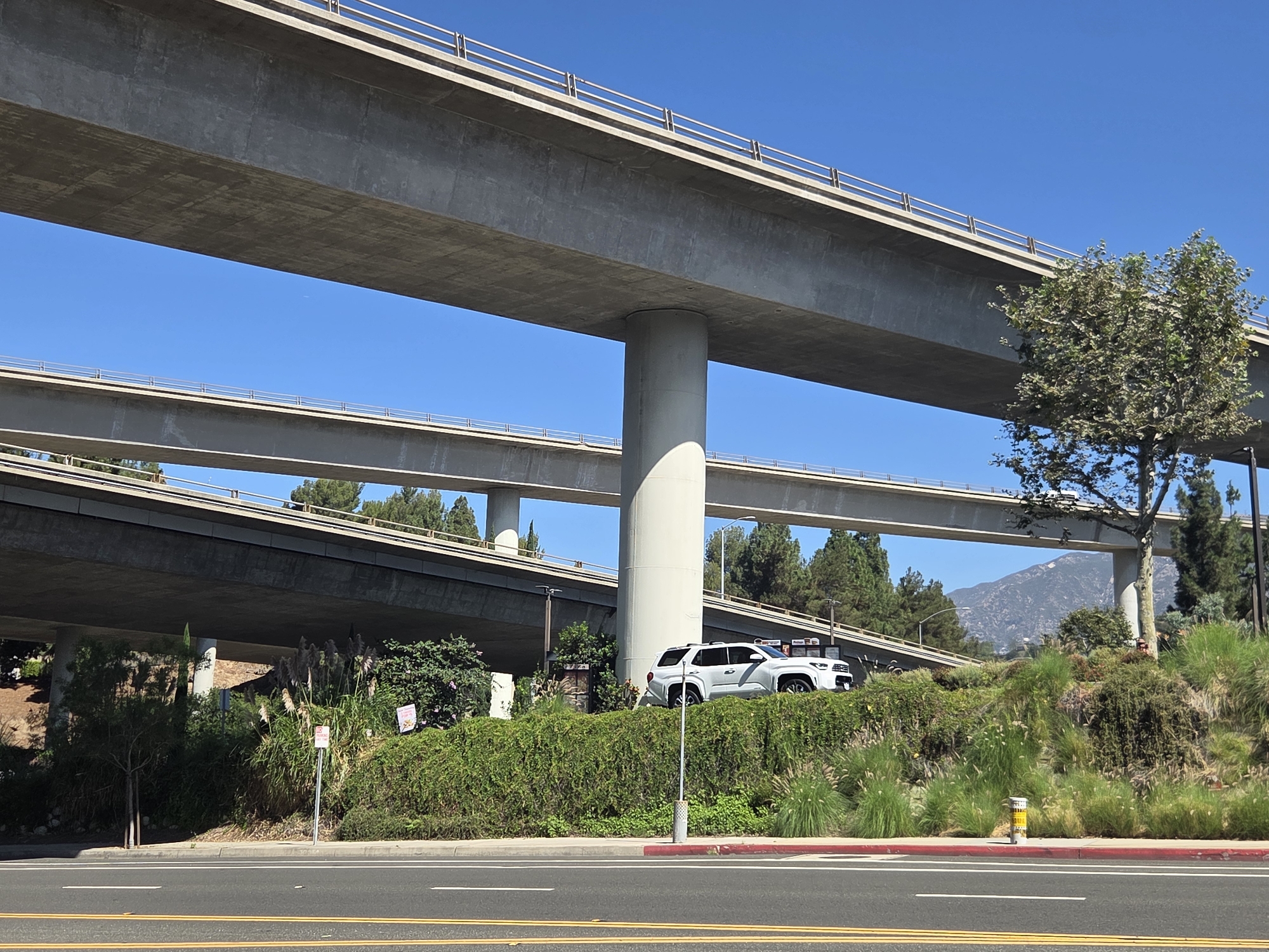 A white SUV is parked on a vegetated slope beneath a multi-tiered concrete overpass in Glendale, California. The SUV, a Toyota Sequoia, is positioned on a grassy area with bushes and tall grasses. Multiple concrete bridges, supported by large cylindrical pillars, dominate the upper portion of the image, creating a layered effect against a clear blue sky. In the background, distant mountains are visible. A paved road with yellow lane markings runs along the bottom of the frame. Several signs are present on the slope, including a "No Parking" sign. The scene appears to be in an urban or suburban environment with extensive road infrastructure.