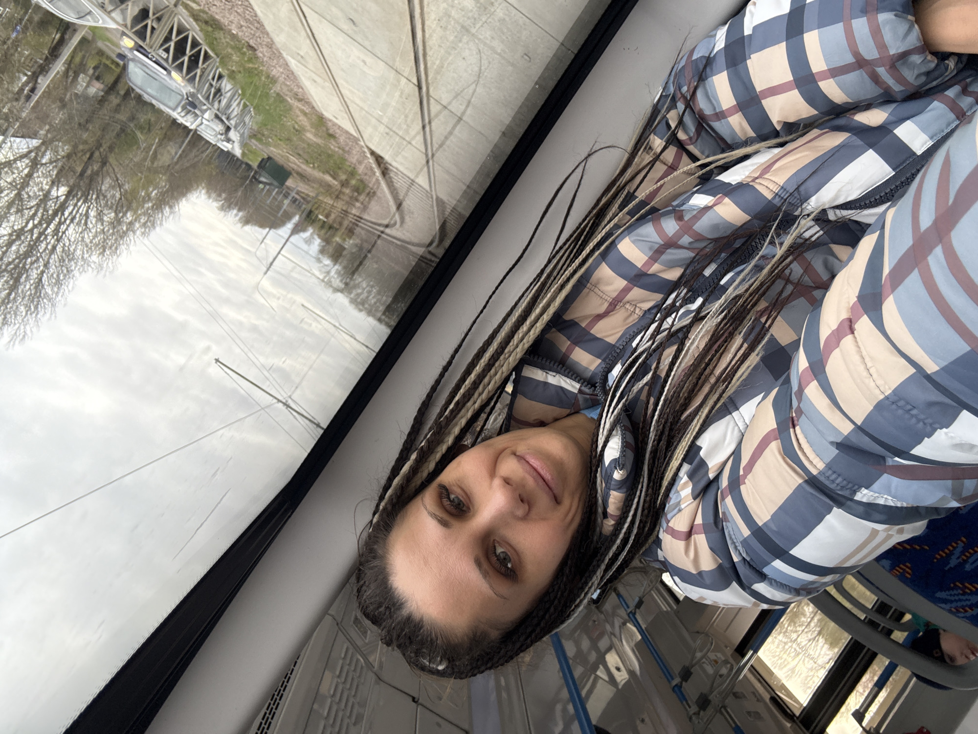 A woman is captured in a selfie-style shot inside what appears to be a public transport vehicle, likely a bus or tram, based on the interior elements and external view. She is positioned in the foreground, looking directly at the camera with a subtle, pleasant smile. Her light-colored eyes are engaging, and her face is framed by numerous long, dark dreadlocks, which feature lighter-toned strands woven throughout.
She is dressed in a puffy, plaid jacket exhibiting a pattern of white, light blue-grey, beige, and reddish-brown/maroon squares. The jacket appears warm and quilted. Behind her, to the right, glimpses of blue patterned seats suggest the interior of a transport vehicle.
To the woman's left, a large window offers a view of the outside world. The sky is overcast and bright, hinting at a cloudy daytime setting. Beyond the window, bare trees dominate the landscape, suggesting a season like late autumn, winter, or early spring. The ground beneath the trees is mostly brown, with some faint patches of green. In the distance, a vehicle, possibly a car or truck, is partially visible, along with some indistinct structures. What appear to be tram or train tracks run parallel to the vehicle, curving away into the distance, reinforcing the context of public transport in motion.
The overall mood is calm, depicting a moment of travel in Rusanovo, Russia. There is no visible text in the image.